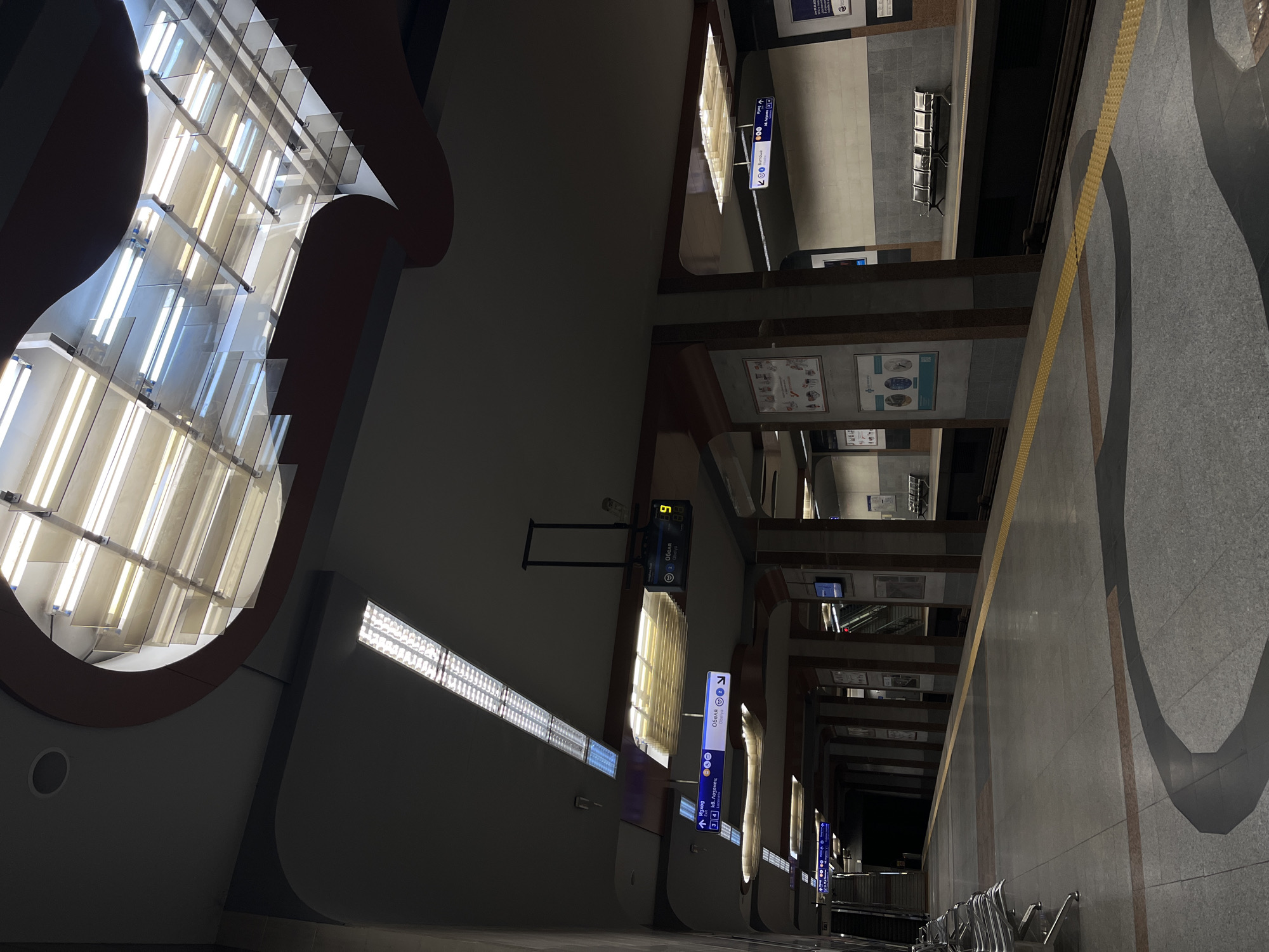 This is an indoor shot of the Sofia Metro station platform in Sofia, Bulgaria. The perspective is tilted, showing the platform edge on the right, a gray stone floor with inlaid designs. Above the platform are overhead structures with signs indicating directions to "Obelya" and "kv. Lozenets," and digital displays showing the time. The left side features a large architectural element with a rounded opening filled with linear lighting. The station design includes a recurring arched motif, visible in the architectural structures and shadows. The lighting is a mix of direct light from the fixtures and ambient light reflecting off the surfaces. The platform appears empty.