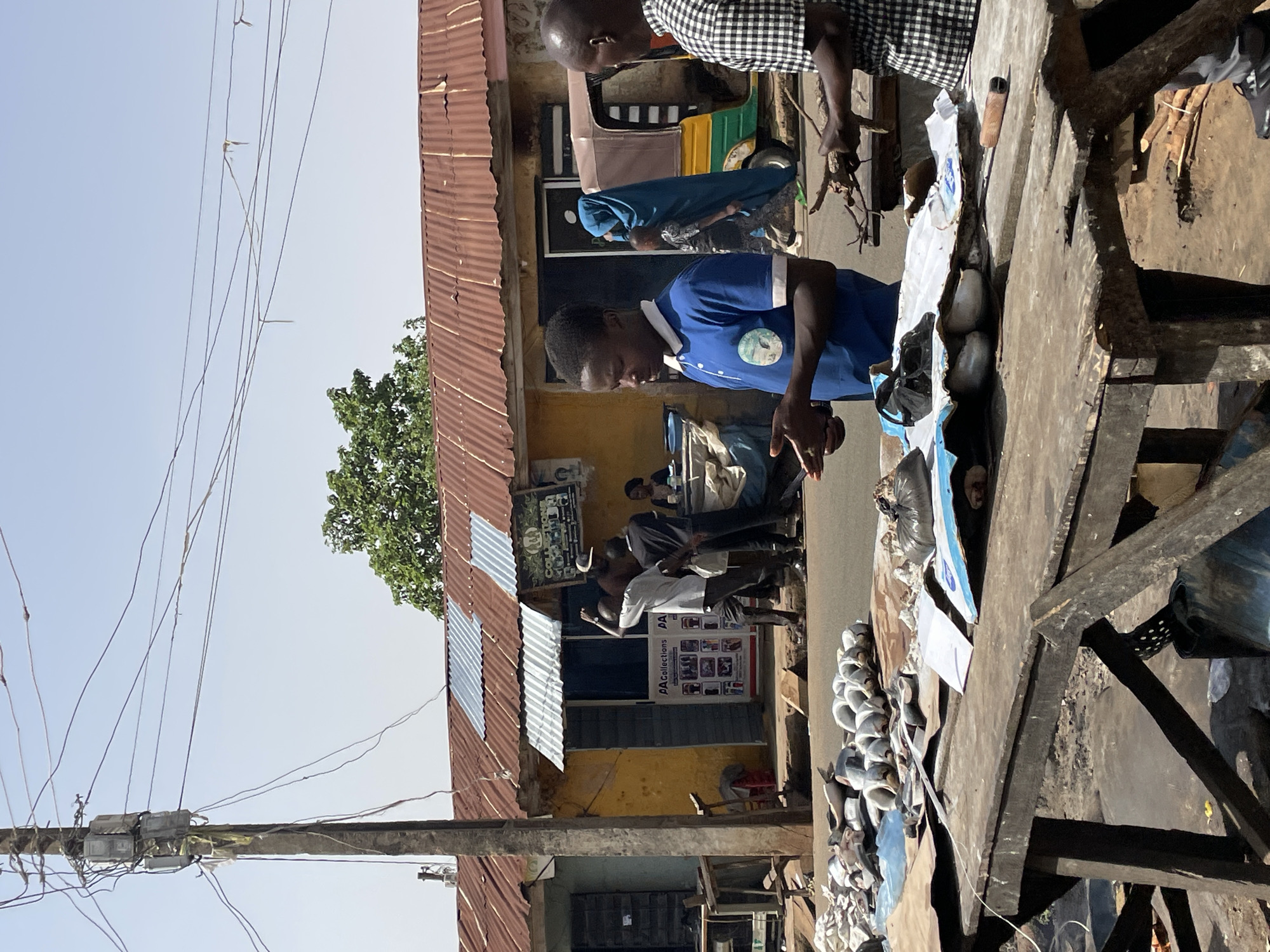 The image shows a street scene in Jos, Nigeria. Two men are working at a wooden table that is overloaded with various items, including round objects (possibly food items) and wrapped packages. One man wears a blue shirt and is focused on something in his hands. The other man, wearing a checkered shirt, appears to be handling sticks or small branches.
Behind the men, there is a yellow building with a corrugated metal roof. A poster is visible on the building’s facade. People are gathered in front of the building. A three-wheeled vehicle is parked nearby. Electrical wires cross the sky above the buildings. The weather appears sunny and clear.