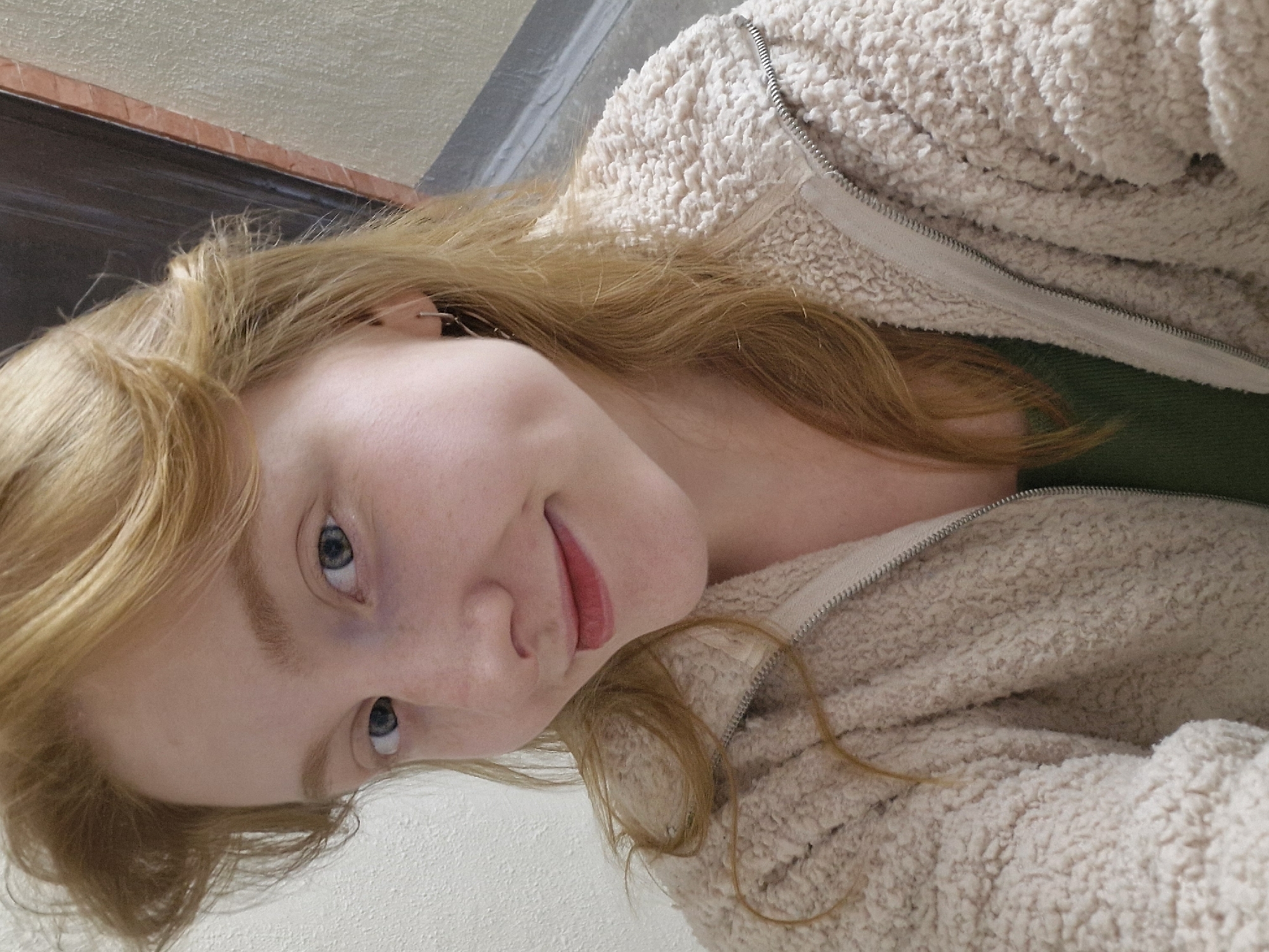 A young woman with blonde hair is the sole subject of this media file. She is wearing a fuzzy, cream-colored hooded jacket over a dark green shirt. Her hair is styled in loose waves and falls around her face. The woman's eyes are blue and she has fair skin with some light freckles. Her lips are painted with a vibrant pink lipstick. She is looking directly at the camera with a slight smile. The background appears to be indoors, with a textured wall on the left and a darker surface above. The lighting suggests it is daytime. No text is visible in the image.