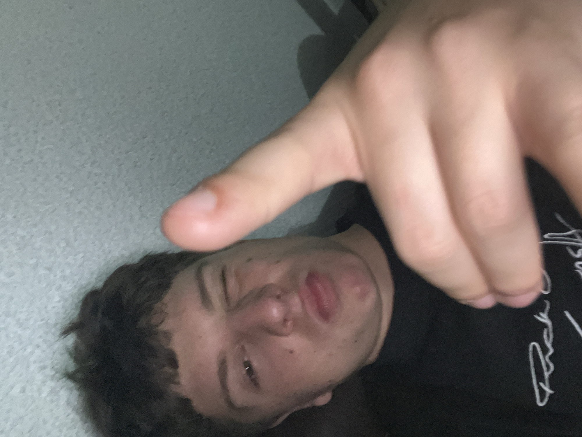 An image depicts a young male with light skin and dark, short, disheveled hair. He is positioned in a close-up, selfie-style view, looking directly towards the camera. The male winks his right eye and slightly puckers his lips. His right hand is extended into the foreground, with the index finger pointed forward, mimicking a "gun" gesture, and the thumb is raised. He is wearing a black t-shirt with white, cursive script lettering visible across the chest, which appears to include the partial word "Rick." The background consists of a uniformly light grey, textured surface, possibly a wall or ceiling, indicative of an indoor setting. The lighting is dim, casting soft shadows.