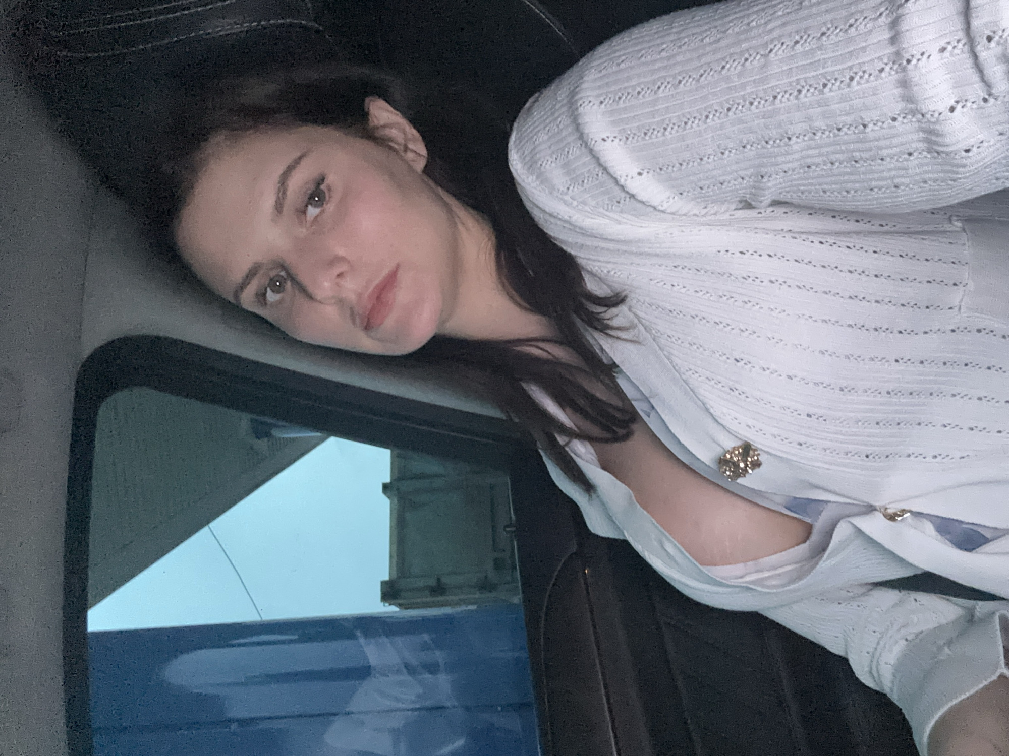 This is a selfie of a young woman, taken indoors, likely in a vehicle. She is lying down, with her head resting on a dark surface, possibly a seat. Her dark hair is spread out behind her. She is wearing a white, open-knit cardigan with decorative buttons, revealing a glimpse of a blue garment underneath. A brooch is attached to the cardigan on her chest.
The woman is looking directly at the camera with a neutral expression. Her skin appears to have a pinkish undertone, and her makeup is subtle.
To her left, a window reveals a cloudy, light-colored sky. Part of a blue object, possibly part of the vehicle's exterior, is visible below the window. The overall lighting suggests it might be daytime, possibly overcast or late in the day. There is no visible text in the image.