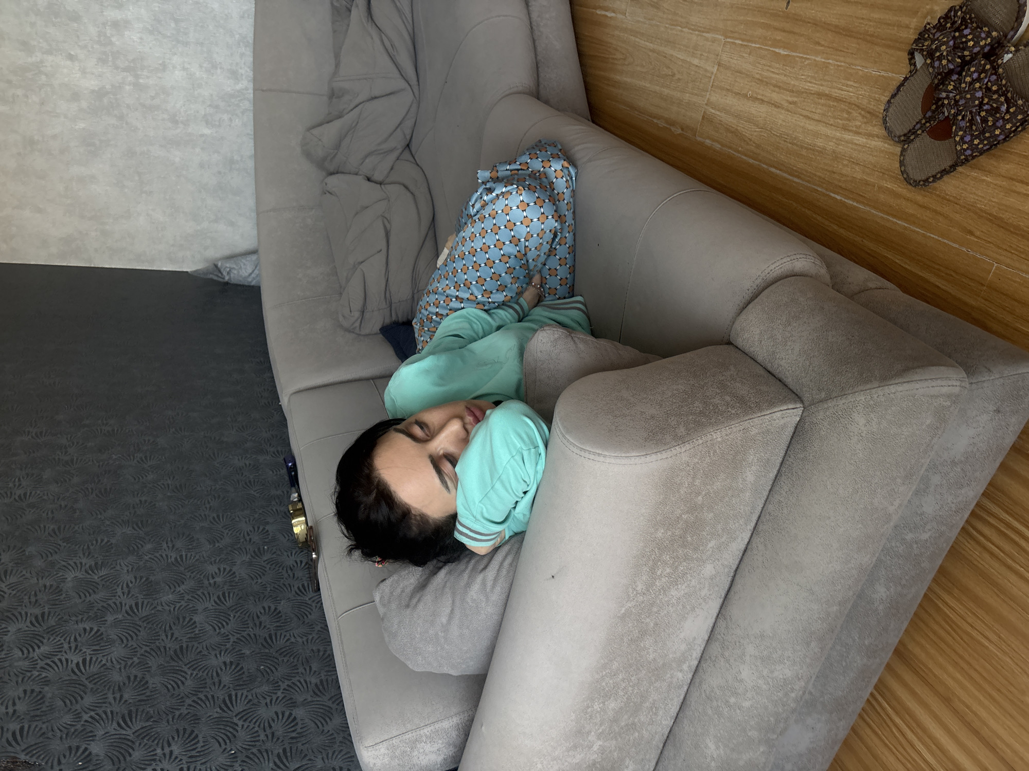 A person lies asleep on a gray sofa in what appears to be an indoor setting. The person is wearing blue patterned pajamas and a light blue sweatshirt. A gray blanket is also on the sofa. To the right of the sofa, there is a wooden wall, with a pair of floral slippers resting on the floor. The room's carpet is dark gray with a subtle, fan-like pattern. The city and specific location within Tbilisi, Georgia, are not discernible from the image.