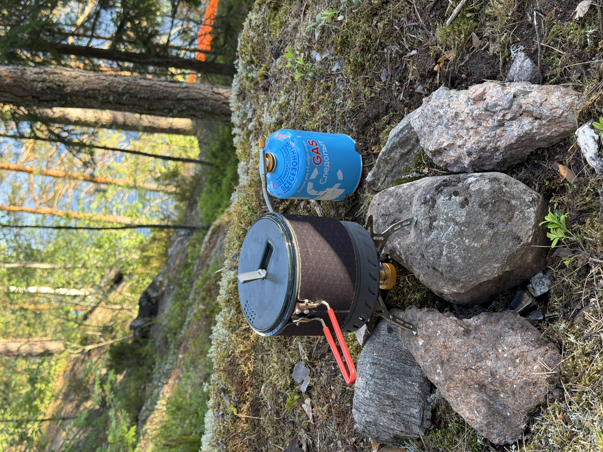 The image depicts a camping stove and a gas canister set up on a mossy, rocky terrain in a forest. The stove, a compact unit with a dark grey pot and a red handle, sits on a small burner. Beside it, a bright blue gas canister with Cyrillic lettering and the word "GAS" in English is connected to the stove via a hose. The background features a sun-dappled forest with tall trees, some with visible bark textures, and a body of water, likely a lake, in the distance. The ground is covered with moss, small green plants, and scattered rocks of varying sizes and textures. The lighting suggests it is daytime, with sunlight filtering through the trees. There are no people visible in the image. The scene is one of an outdoor campsite or a picnic spot within a natural environment. The location is identified as Oyayarvi, Russia.