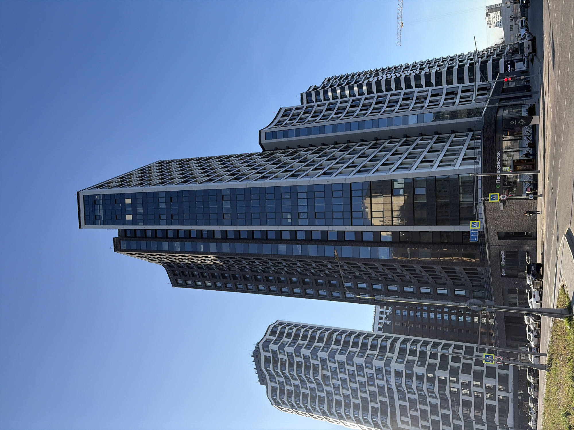 The media file depicts a modern urban scene in Moscow, Russia, under a clear blue sky, suggesting a sunny day. The primary subjects are a series of contemporary, multi-story buildings with distinct architectural styles. One prominent building features a wavy, undulating facade with a pattern of alternating dark windows and white, protruding elements. Other buildings exhibit a more rectilinear design with extensive glass facades and repeating patterns of balconies or window sections.
In the foreground, a street runs alongside the buildings, with parked cars visible. Pedestrians are also present, one walking on the sidewalk and another near a crosswalk. Traffic lights and street signs are visible, indicating an active urban environment. One sign displays Russian text, "Перекрёсток" which translates to "Crossroads" and also a store name.
The time of day appears to be daytime, likely late morning or early afternoon, given the bright sunlight casting shadows. The overall atmosphere is one of a developing or developed urban area, characterized by modern architecture and everyday city life. The image captures a dynamic cityscape, showcasing the blend of residential and commercial spaces typical of a large metropolis. The presence of a construction crane in the background suggests ongoing development or construction in the area.