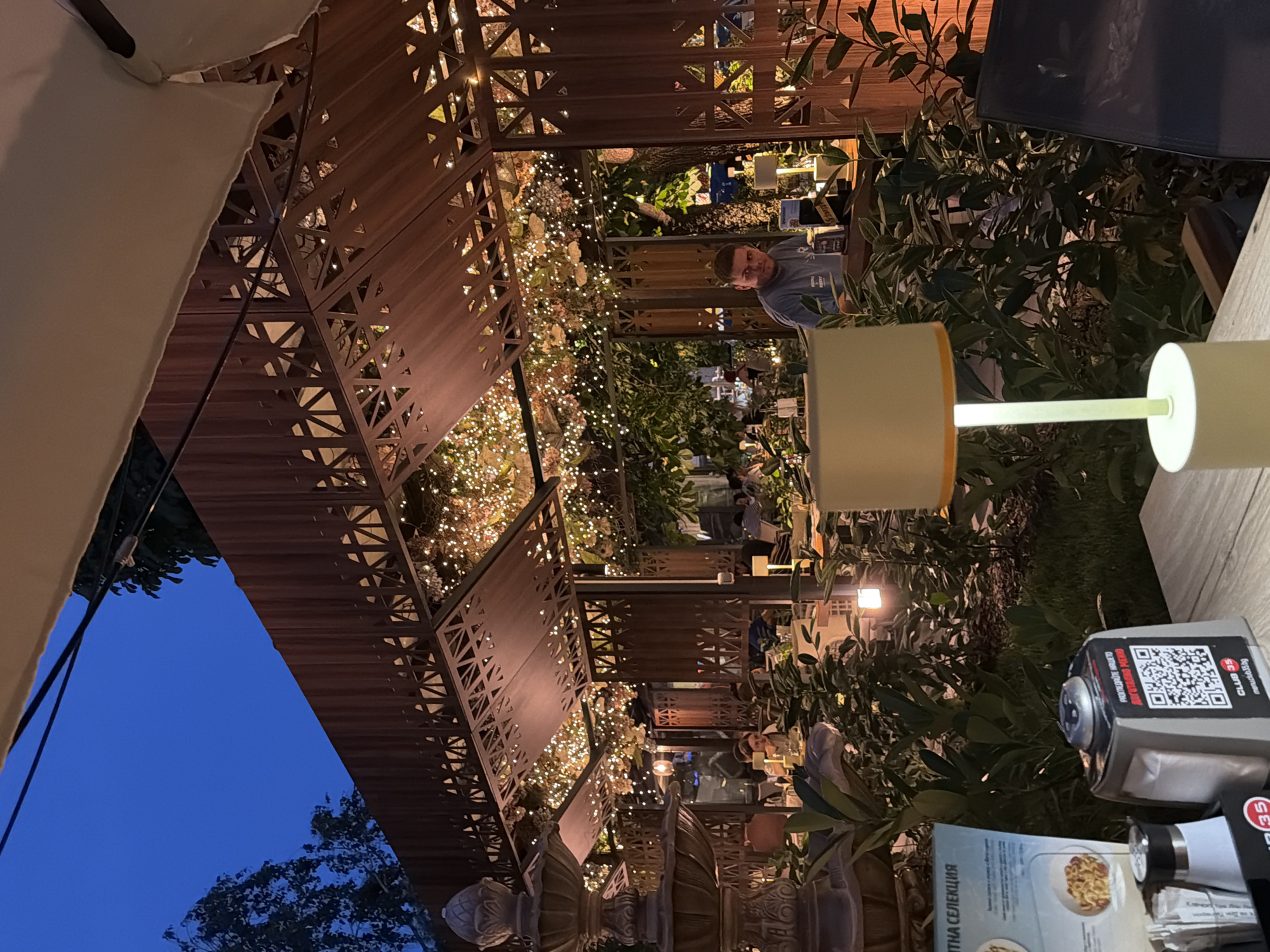 The image depicts an outdoor seating area of a restaurant or cafe at dusk in Sofia, Bulgaria. The establishment features a wooden structure with intricate geometric cutouts, draped with numerous small string lights, creating a warm and inviting ambiance. Lush green foliage surrounds the seating area, with a prominent fountain visible on the left side of the frame.
In the foreground, a table is set with a lamp with a light yellow shade, a salt and pepper shaker, and a napkin dispenser with a QR code that reads "CLUB 35 menuc35.bg." A menu with images of pasta dishes is also visible.
In the middle ground, several people are seated at tables, appearing to be dining or socializing. One man in a blue t-shirt is looking directly at the camera. Further back, other patrons are visible, some interacting with each other, others engaged in their meals.
The overall scene suggests a relaxed evening at a well-lit outdoor dining venue, likely popular for its atmosphere and setting. The presence of specific branding on the QR code and menu suggests a commercial establishment.