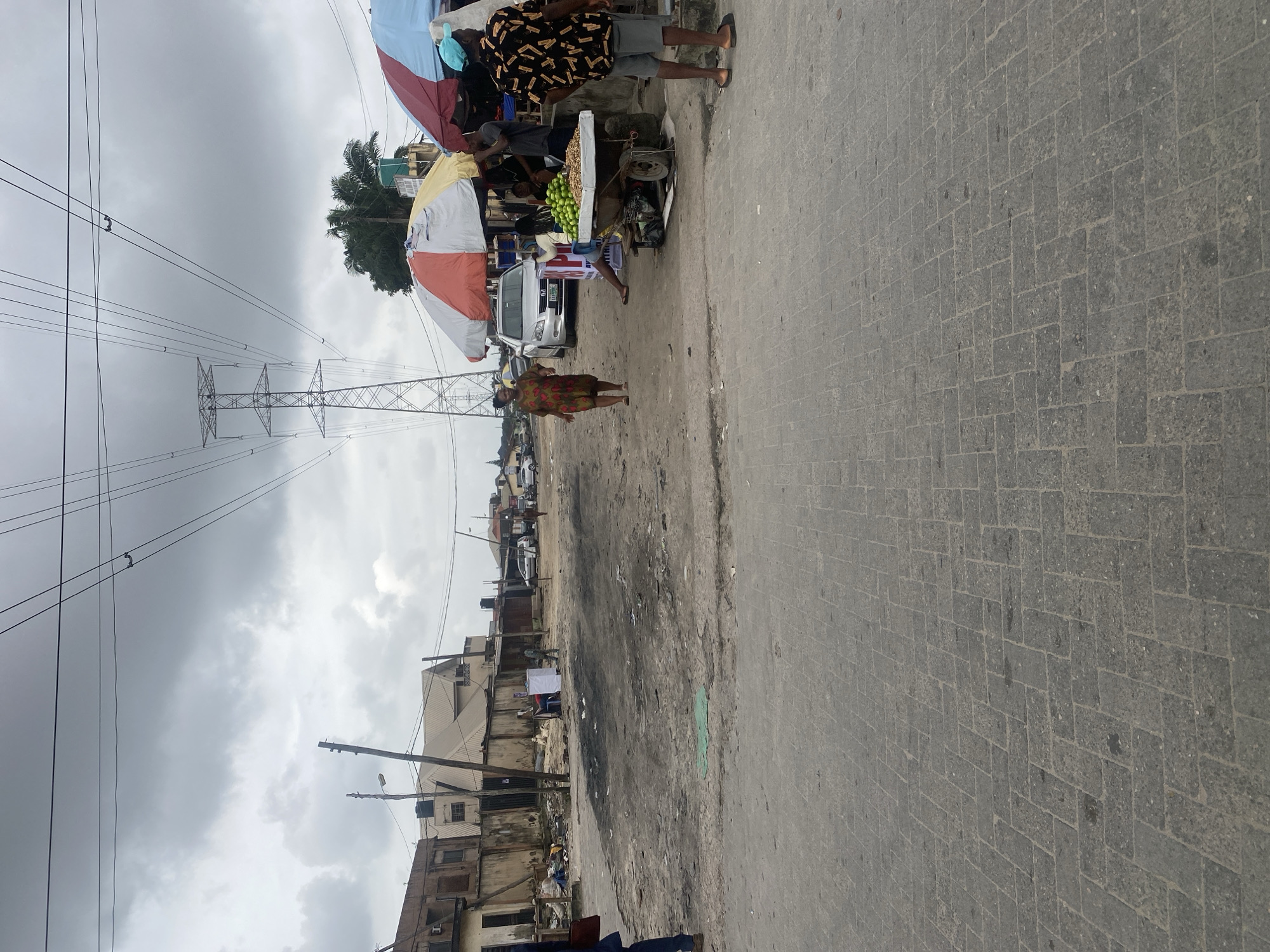 A street scene in what appears to be an urban environment under a cloudy sky. In the foreground, a paved road with a cobblestone pattern leads towards the background. A dirt and debris-filled area is visible to the left of the road. On the right side of the street, there is a market stall with colorful umbrellas, selling produce, primarily what appear to be limes. Several people are present. One woman in a patterned dress walks across the street, while others are gathered at the market stall. In the background, there are buildings, some appearing weathered, and a large electrical tower with power lines stretching across the sky. A silver sedan is parked near the market stall. The overall impression is of a busy, functional street with commercial activity.