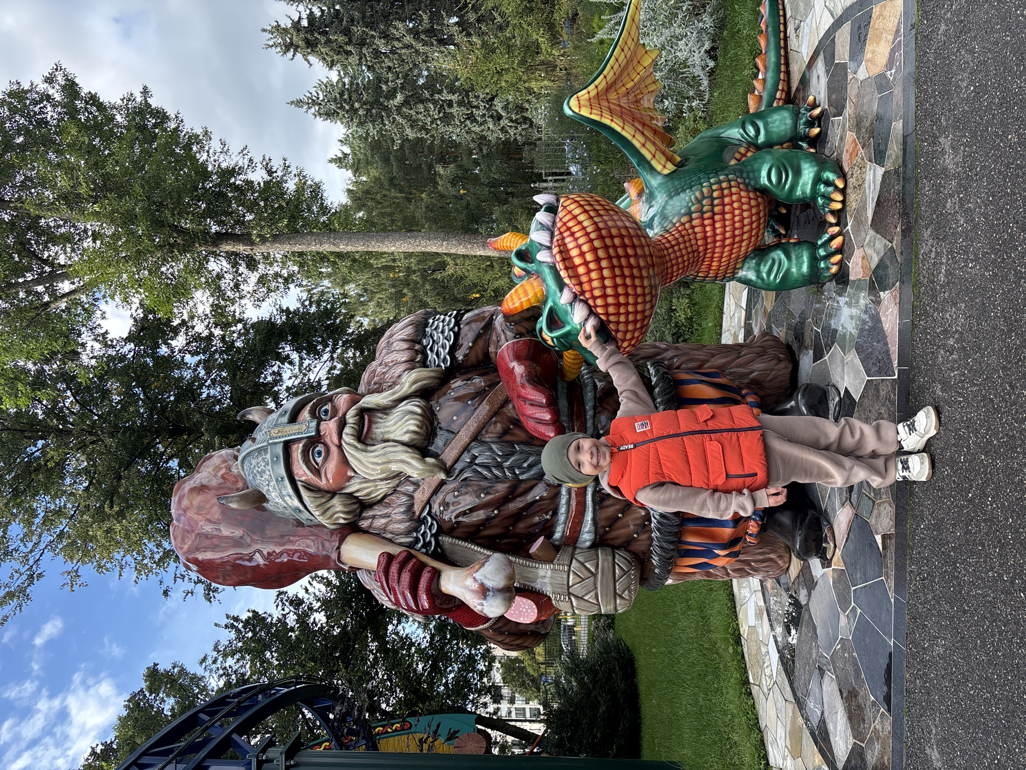 A young boy in a red vest and grey sweatpants stands between a large statue of a Viking and a statue of a green dragon in an outdoor park. The boy is smiling and touching the dragon's snout. The Viking statue is larger than the boy and is holding a large piece of meat over its shoulder. The dragon statue is green with orange and yellow accents, and has wings. The ground in front of the statues is paved with irregular grey, black, and brown stones. Behind the statues, there is green grass, trees, and a blue sky with clouds. A rollercoaster track is visible in the background on the left side of the image. The setting appears to be an amusement park or a themed area within a park.