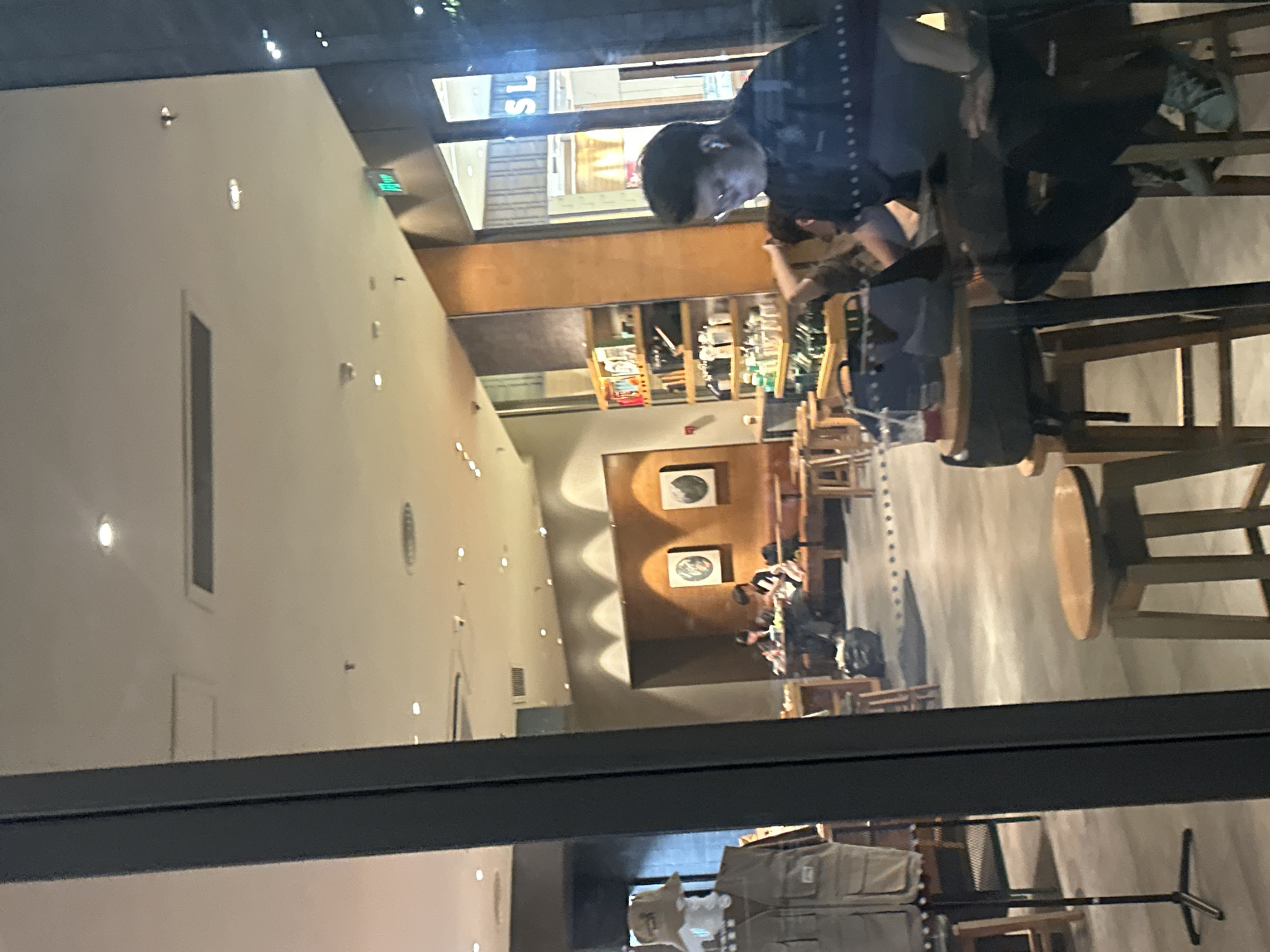 The image depicts the interior of a cafe, possibly a Starbucks, viewed from outside through a window. In the foreground, to the left, a mannequin dressed in a tan vest and a bucket hat is displayed. Further inside the cafe, several people are seated at tables. Two individuals are visible at a table towards the center-left, one appears to be writing. Towards the right of the frame, a person wearing glasses and earbuds is seated at a table, looking down at a laptop. The cafe's interior features wooden tables and chairs, shelves stocked with merchandise, and wall art. The lighting is warm, with overhead lights and accent lighting along the walls. The overall scene suggests a casual, relaxed atmosphere. The specific location in Chongqing High-tech Zone, China cannot be determined from the visual information provided in the image.