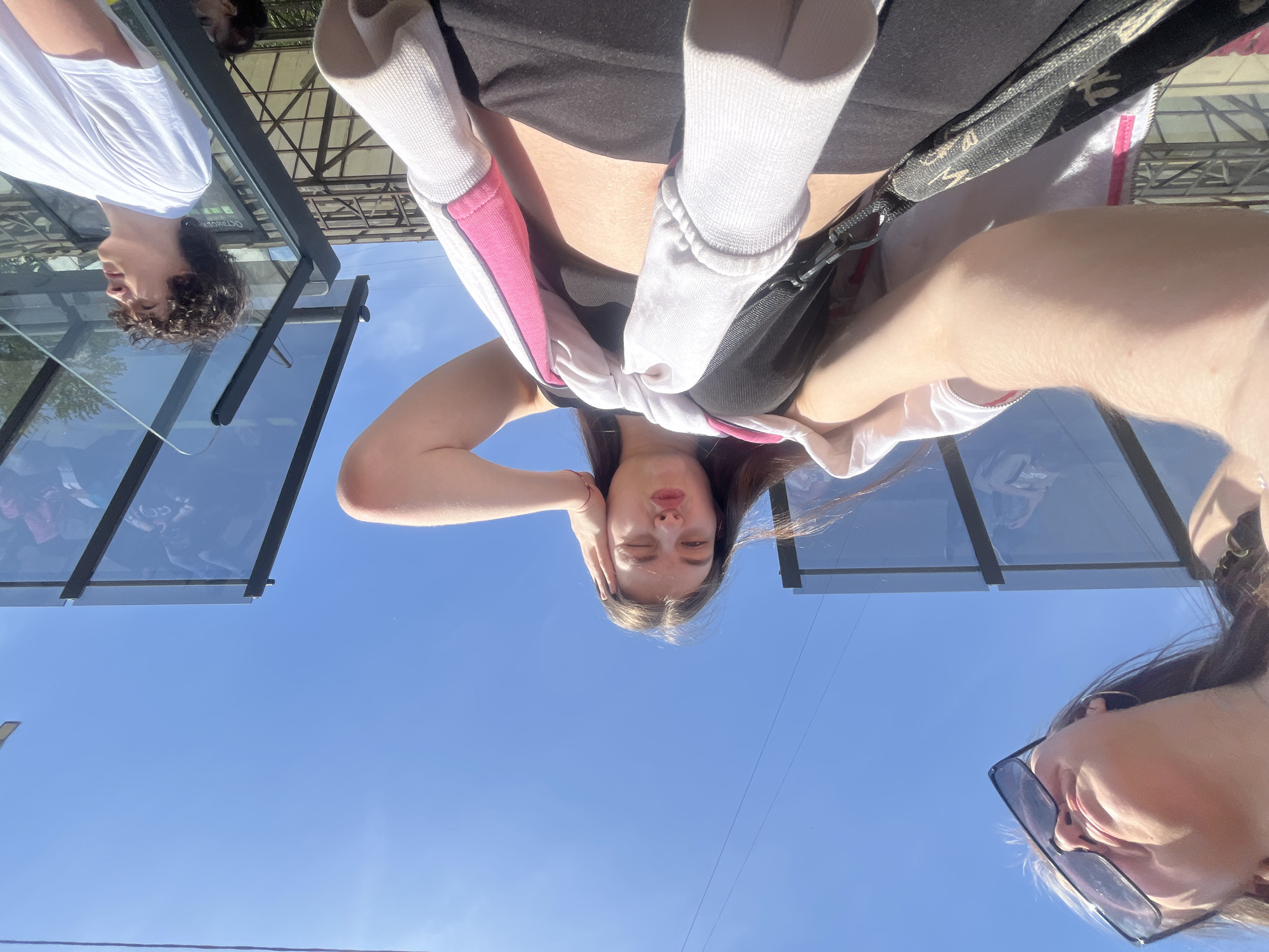 From an upside-down perspective, the image shows a group of people gathered outside near what appears to be a bus or tram stop shelter. The sky is clear and blue. Three individuals are partially visible. One person is wearing a white shirt and has curly hair. Another person is wearing a black top, a pink and white sweatshirt, and a bag. A third person is wearing sunglasses. The location is likely Saint Petersburg, Russia, based on the Cyrillic text visible on the shelter. The general setting suggests an urban environment.