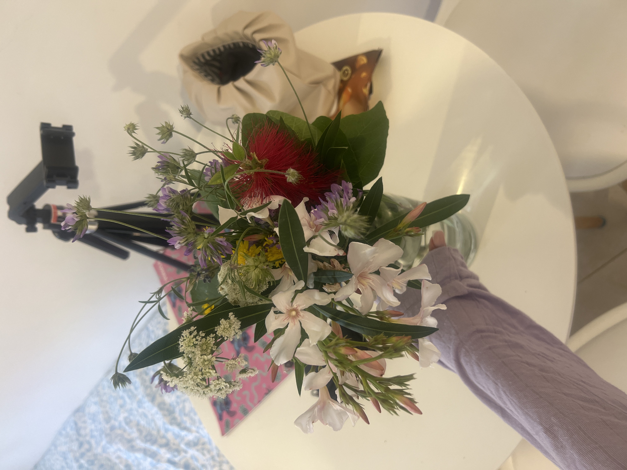 A still life image shows a bouquet of mixed wildflowers in a glass vase placed on a white, round table. The bouquet includes white oleander blooms, a red bottlebrush flower, and various other purple, yellow, and white wildflowers, with greenery. To the left of the vase, a black tripod with a phone mount is positioned on the table. A beige tote bag and a brown package sit in the background, slightly out of focus. The table appears to be positioned in a room with tiled flooring and white chairs. A person's arm wearing a light purple long-sleeved shirt extends toward the vase. The location is indicated to be in Budva, Montenegro.