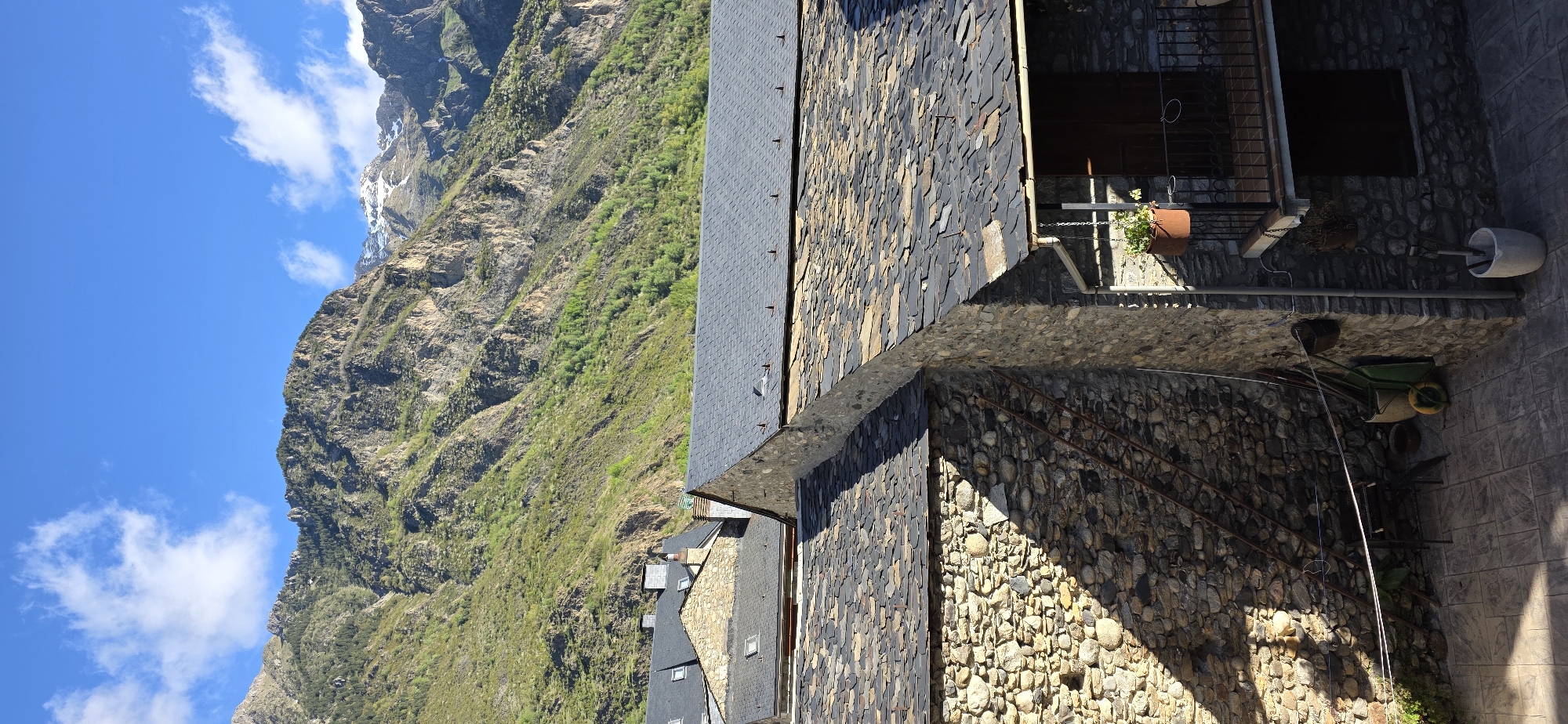 The image depicts a scene in the city of Benasque, Spain. In the foreground, a stone building with a dark tiled roof is prominently featured, alongside a stone balcony adorned with a potted plant. A wheelbarrow is visible in the immediate foreground. The backdrop showcases a mountain range, with some snow cover on the higher peaks, set against a clear blue sky with scattered clouds. The steep slope of the mountain is covered with a mixture of rock and greenery. The overall setting conveys an outdoor, mountain village environment.