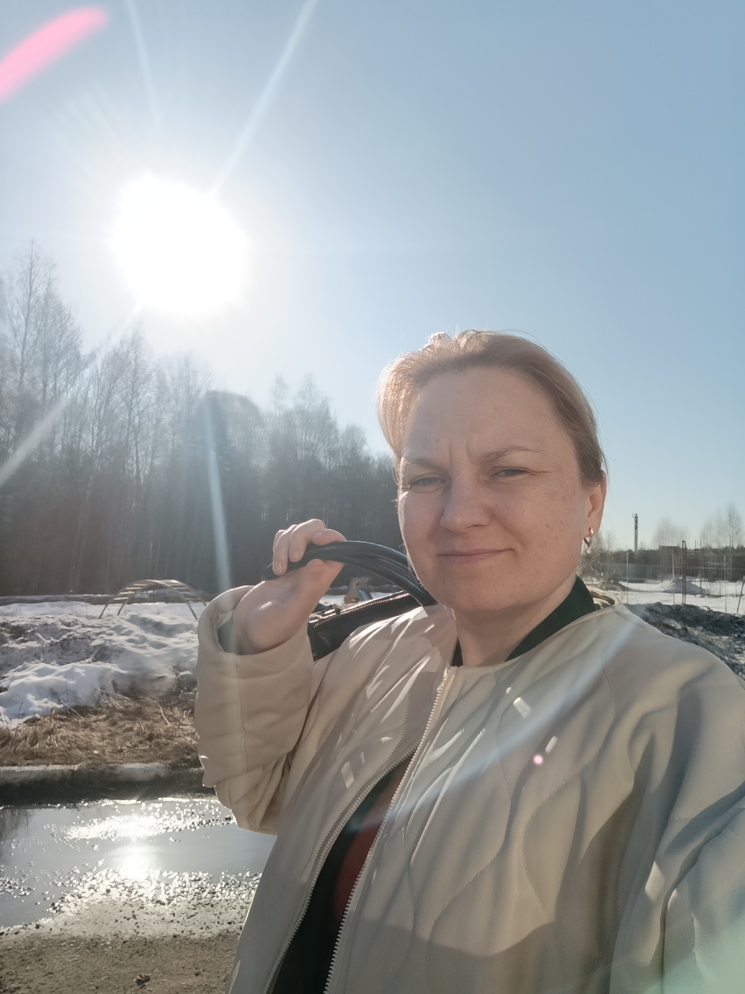 A woman with fair skin and light brown hair is seen in an outdoor selfie, wearing a light-colored quilted jacket. She is holding a black object over her shoulder. The background shows a landscape in what appears to be late winter or early spring, with patches of snow and bare trees. There's water in the foreground, reflecting sunlight. The bright sun is positioned high in the sky, partially obscured by the trees. The location is in Марковский, Russia.