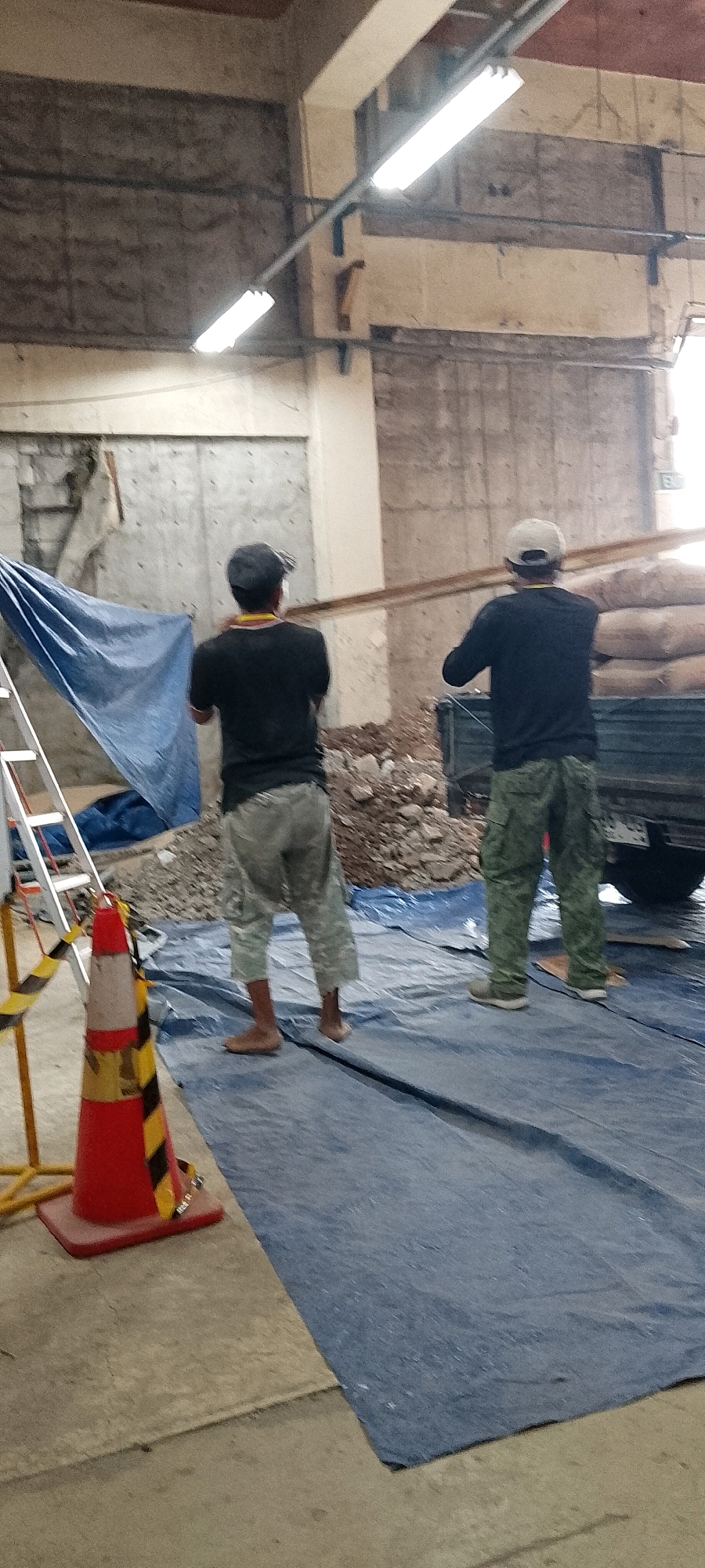 Two men are working in a construction or warehouse environment in Depok, Indonesia. The man on the left is wearing a black t-shirt, camouflage pants, and a cap, carrying a plank of wood on his shoulder. The man on the right, also wearing a black shirt, camouflage pants, and a white cap, is assisting him. They are on a blue tarp laid on the ground. In the background, there is a pile of rubble, a truck with sacks, and a ladder. The scene appears to be taking place indoors, possibly in a partially demolished or under-construction building, as suggested by the concrete walls and exposed ceiling beams with fluorescent lighting. A red and yellow traffic cone is visible in the foreground, indicating a work zone or safety precaution.