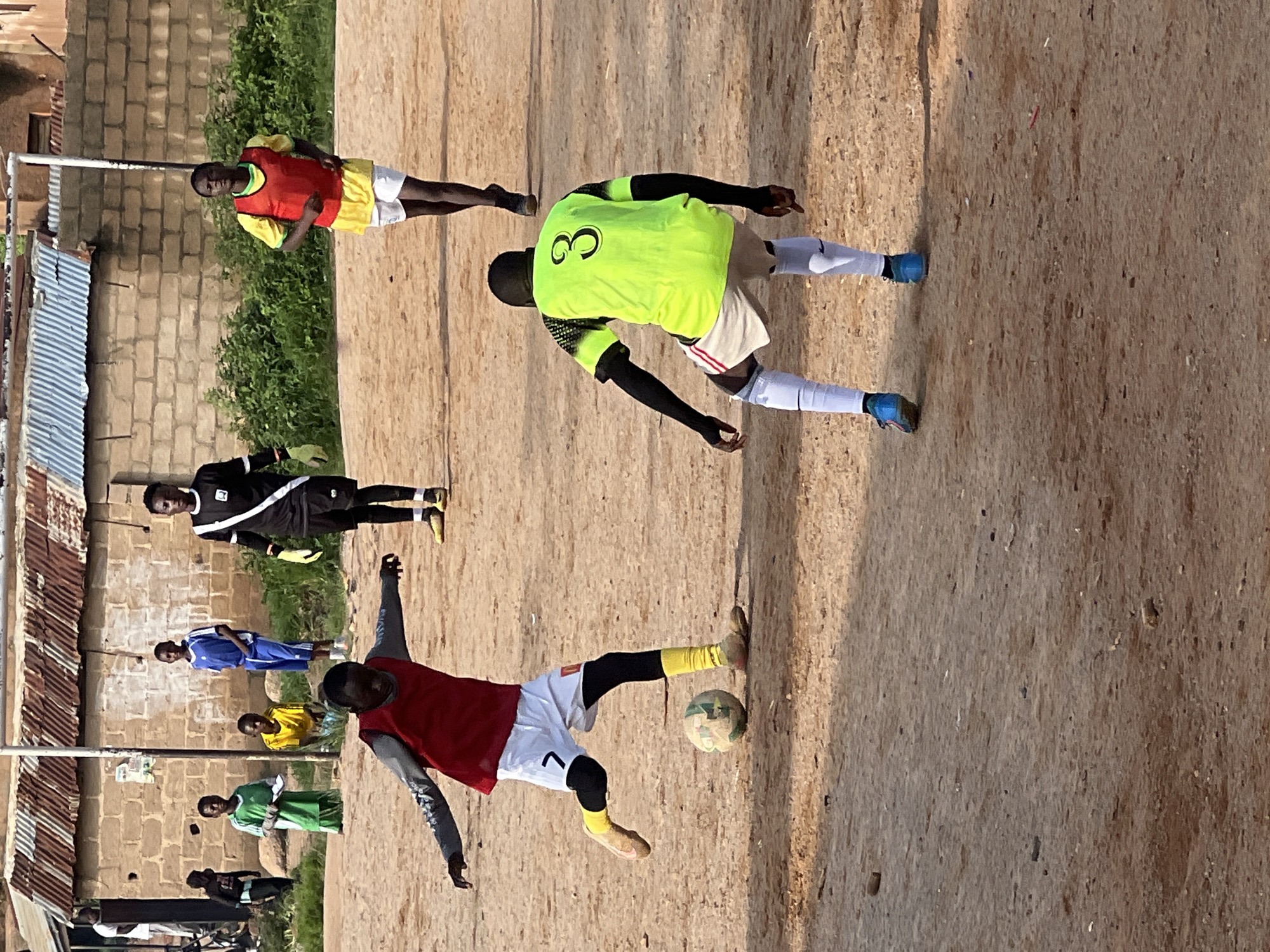 The image depicts a football game being played on a dirt field, likely in Jos, Nigeria. In the foreground, two young men are actively engaged in the game. One, wearing a red vest over a gray long-sleeved shirt and white shorts with the number 7 visible, is kicking a soccer ball. He has yellow socks and black leggings. The other, in a bright lime green short-sleeved shirt with the number 3 on the back, black sleeves, and white shorts with red trim, is positioned as if to receive or defend. He is also wearing white socks and blue cleats.
In the background, several other individuals are present, observing or participating in the game. A person in a black goalkeeper jersey with white accents and gloves stands in the middle ground. To their left, a person in a yellow shirt and red vest is visible. Further to the left, a young person in a green shirt and shorts, and another in a blue soccer kit, are also present. Behind them, a brick building with a corrugated metal roof, typical of some structures in Jos, can be seen. The environment is dusty and dry, characteristic of the terrain in Jos, particularly during the dry season. The overall scene captures a moment of amateur youth football in a developing urban setting.