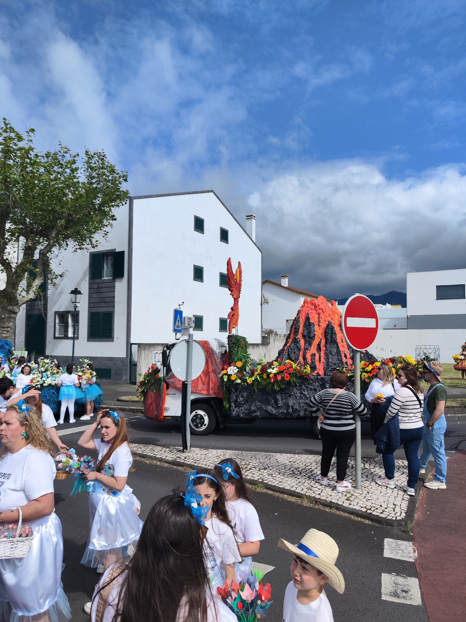 The image captures a street scene in Ribeira Grande, Portugal, likely during a festive event or parade. A large float resembling a volcano, complete with "lava" and floral decorations, is positioned on a flatbed truck. A "No Entry" traffic sign stands in front of it. The float is partially obscured by people, including women in matching white outfits with blue trim, possibly participants in the parade. A child wearing a straw hat with a blue ribbon is also present. Spectators stand on the sidewalk, some observing the float. Background buildings consist of white structures with dark-framed windows. The sky is partly cloudy. A tree with green foliage is visible on the left.