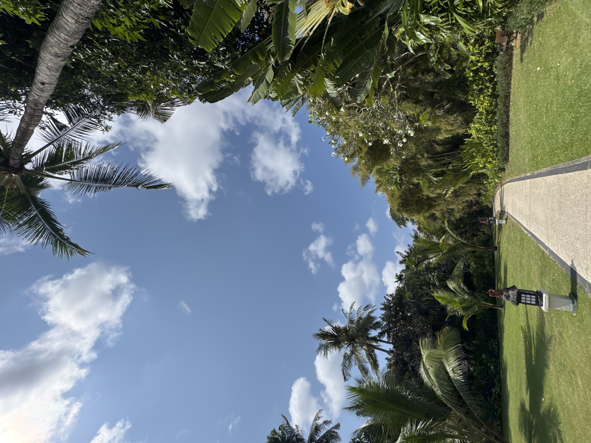 The image shows a paved pathway curving through a lush green lawn in a tropical setting. Tall palm trees and dense foliage frame the path, with a clear blue sky dotted with white clouds overhead. Several decorative lanterns with a rustic design are placed along the left side of the pathway. In the distance, more palm trees and various tropical plants are visible. The overall scene suggests a well-maintained garden or resort area in a warm climate, likely Kuta Selatan, Indonesia, known for its tropical landscapes and resorts.