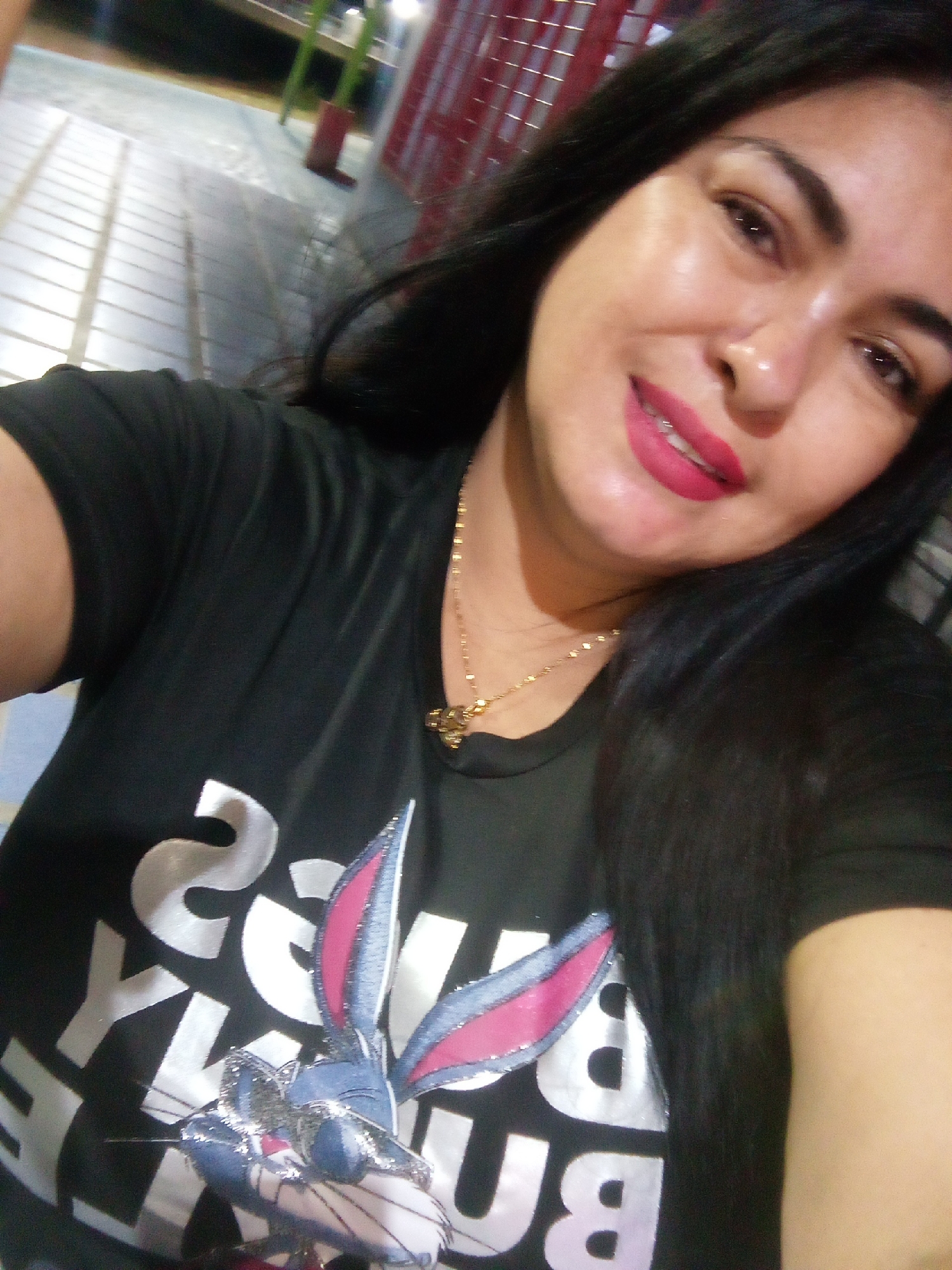 A woman is in an unknown city, wearing a black t-shirt with a Bugs Bunny graphic. She is wearing lipstick and a necklace. The background is a blurred cityscape.