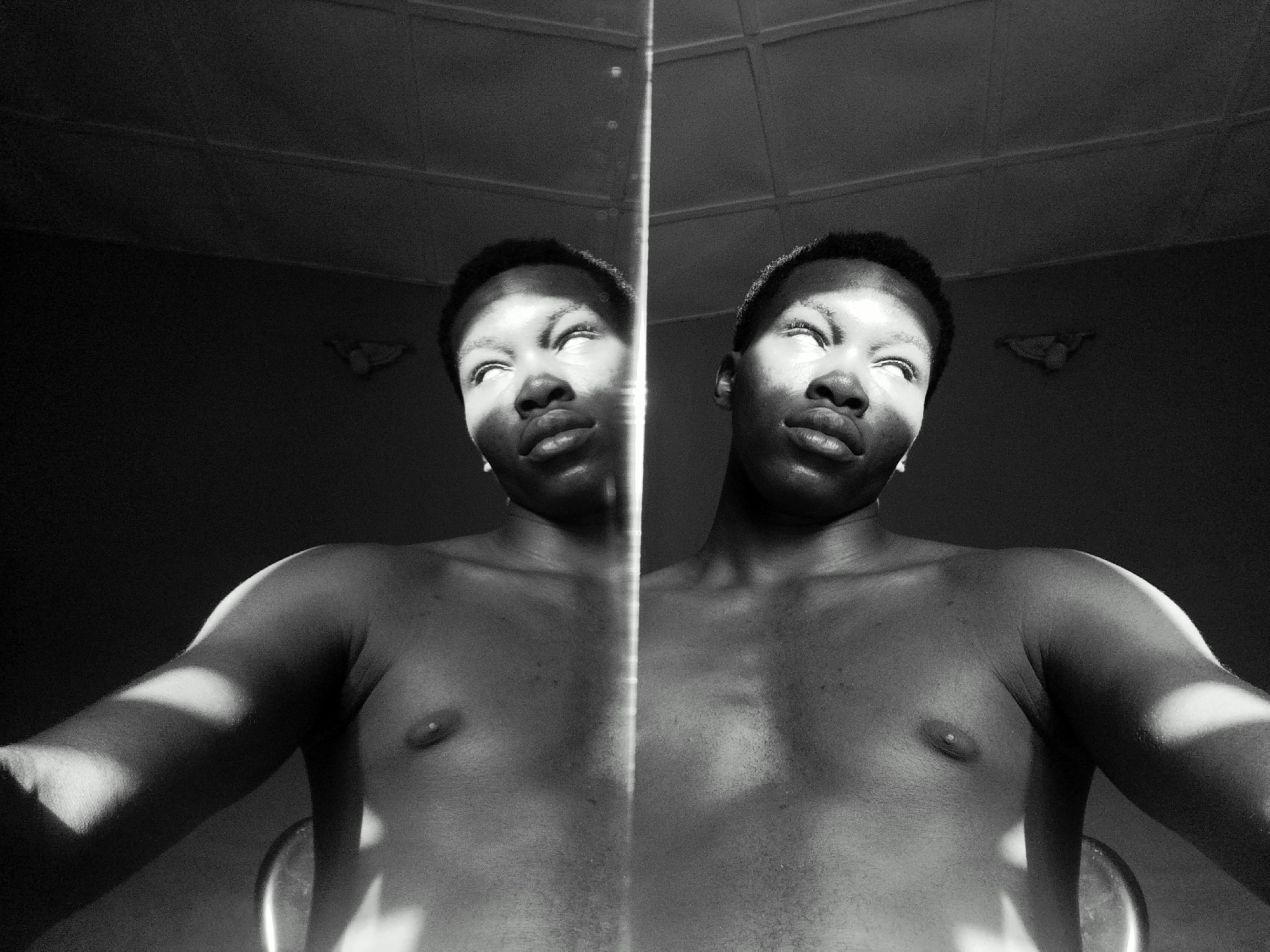 A black and white photograph captures a shirtless dark-skinned man reflected in a mirror, creating a double image. Both the man and his reflection are looking upwards with their eyes slightly rolled back. A sharp vertical line dissects the image, marking the edge of the mirror. Light streaks across his face, chest, and arms. The background is dark and indistinct, revealing a ceiling with rectangular panels. The location appears to be indoors in the city of Keffi, Nigeria.