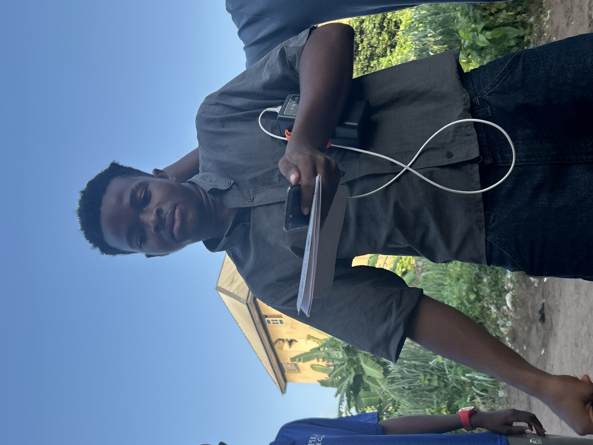 The image depicts an outdoor scene in Ihiagwa, Nigeria, under a clear blue sky. A young Black male, the central figure, is shown from the chest up, looking directly at the camera. He is wearing a dark grey button-down shirt and dark denim jeans. He is holding a smartphone in his left hand and a stack of papers in his right. Attached to his shirt on the right side is a black portable power bank, connected by a white cable that loops in front of him. To his left, another young Black male is partially visible, wearing a blue t-shirt and a red watch on his left wrist. To the right of the central figure, a third individual is partially visible, wearing a blue shirt. In the background, a light yellow, two-story building with a peaked roof is visible, surrounded by lush green vegetation, including banana trees. The ground in the foreground appears to be dirt with some scattered debris.