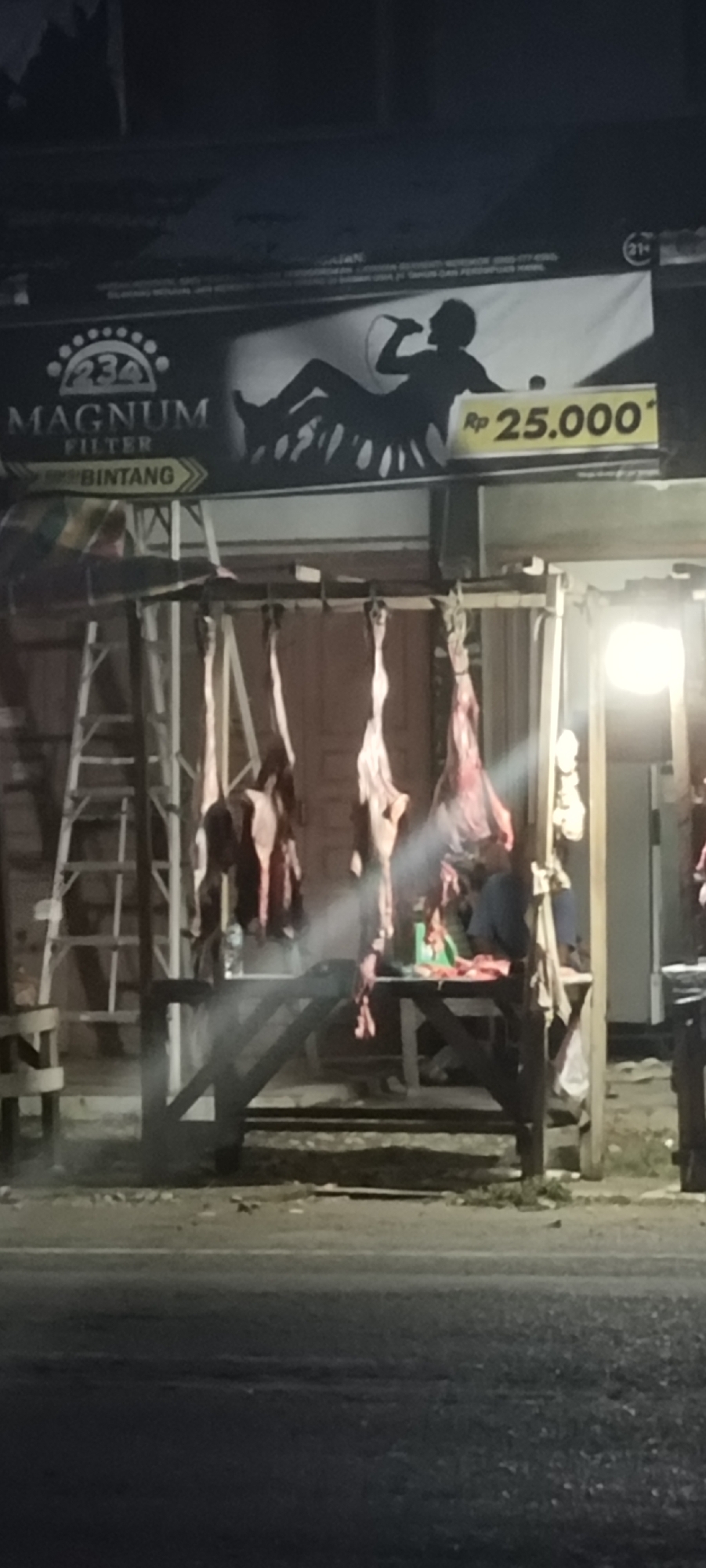 The scene captures a nighttime street view in Lhokseumawe, Indonesia, showcasing a roadside meat stall illuminated by artificial light. Hanging from a wooden frame are multiple animal carcasses, seemingly beef, with portions of the hide still attached. Behind the carcasses, a person stands at a table, possibly preparing the meat. A ladder leans against the side of the structure. Above the stall, a billboard advertises "Magnum Filter" and "Bintang," with a silhouette of a person holding a microphone and the price "Rp 25.000" displayed. The foreground shows a street surface.