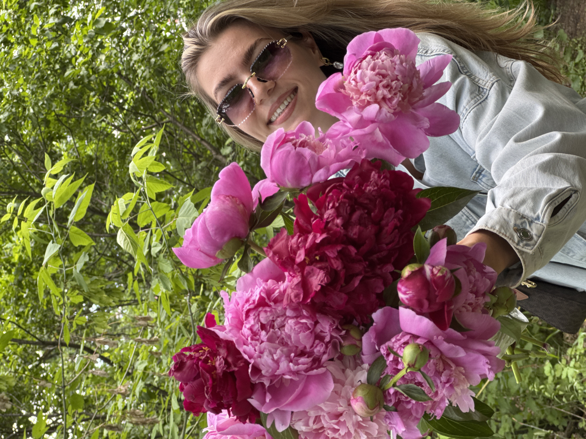 A young woman is captured from a low-angle perspective, smiling broadly while holding a large, abundant bouquet of peonies. Her long, light-colored hair flows around her shoulders. She wears fashionable, rimless sunglasses with small decorative elements on the frames and a light blue denim jacket. Her bright smile reveals her teeth, conveying a sense of happiness and warmth.
The bouquet dominates the lower and central part of the frame, showcasing numerous large, full-bloom peonies. The flowers exhibit a rich spectrum of pinks, fuchsias, and deep reds, with some unopened buds also visible, suggesting freshness. The woman holds the flowers up, seemingly presenting them or posing for the camera.
The setting is outdoors, with a dense background of lush green foliage and tree branches, indicating a garden, park, or natural environment. The bright, natural illumination suggests it is daytime. The overall ambiance of the image is cheerful and vibrant, highlighting the woman's joy and the beauty of the flowers. There is no visible text in the image.