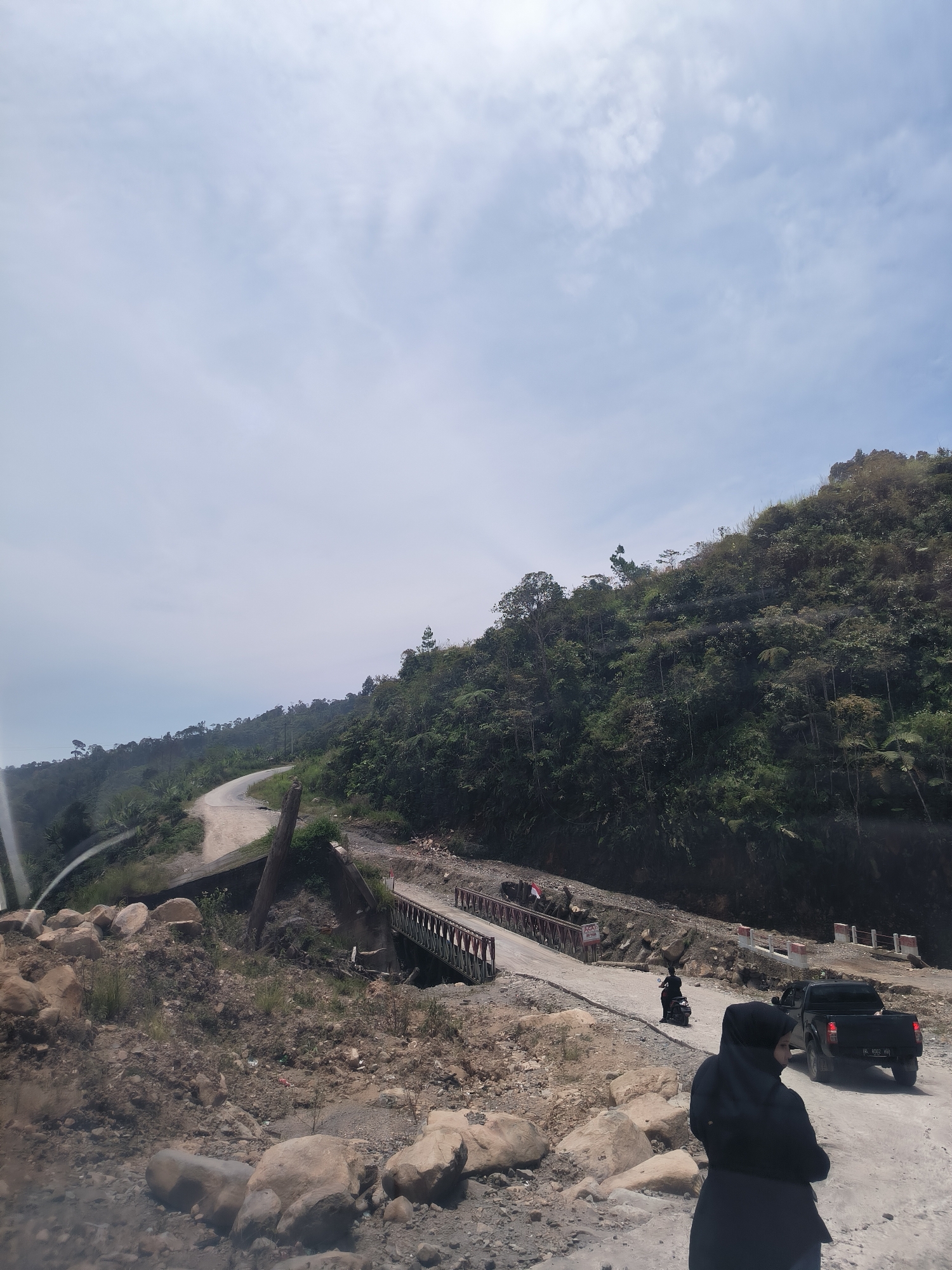 A winding, unpaved road traverses a lush, green mountainside under a bright, cloudy sky in Bintangpermata, Indonesia. The road is flanked by dense vegetation and rocky terrain. Several vehicles are visible: a black pickup truck, a motorcycle with a rider, and another motorcycle further up the road. A small, red and white bridge crosses a gap in the road, with signs and barriers along its edges. In the foreground, a person wearing a black hijab stands with their back to the camera, looking towards the road. The overall scene depicts a rural, undeveloped area with ongoing road construction or maintenance.