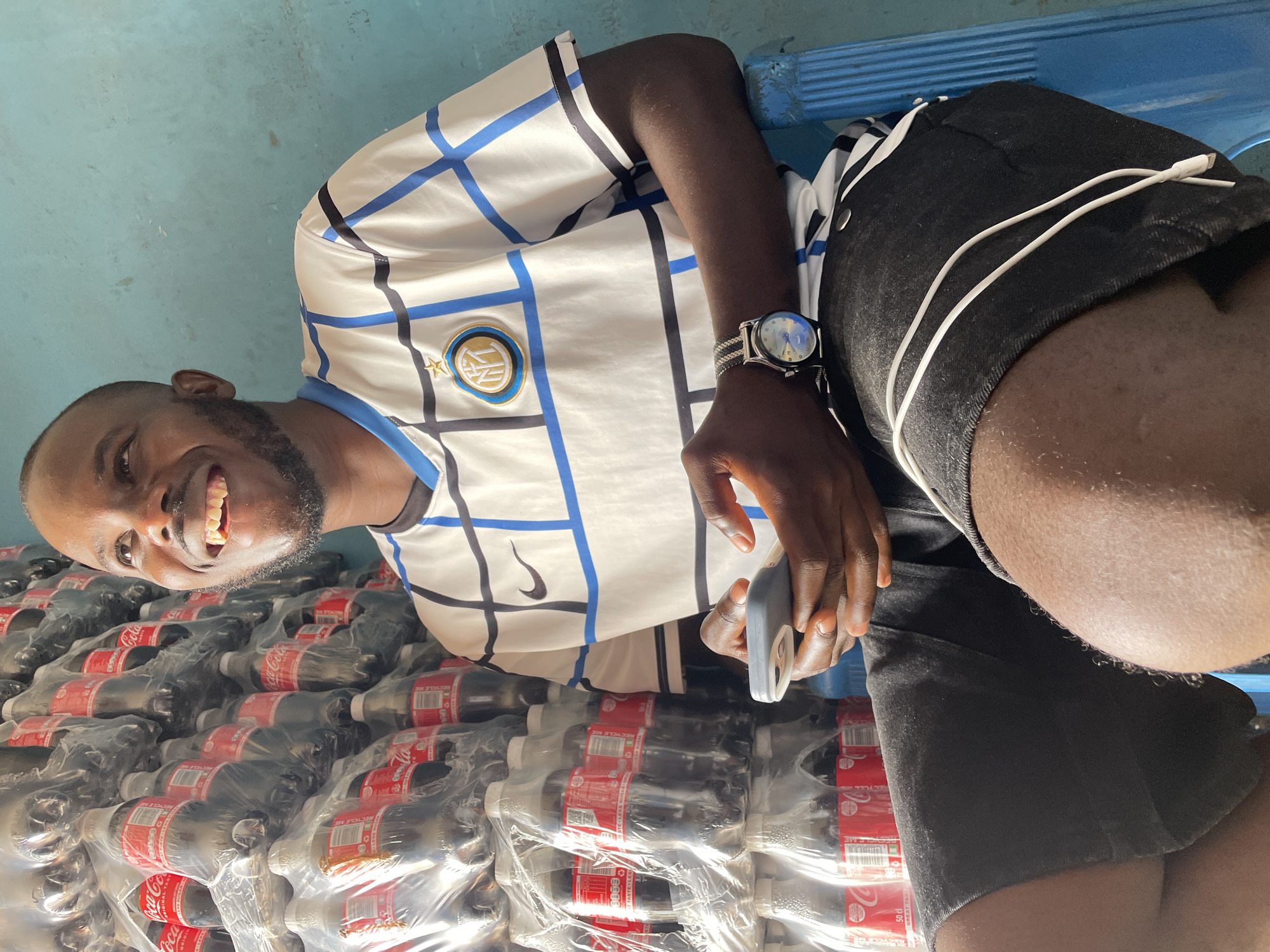 The image features a dark-skinned man in a white Inter Milan jersey with blue and black accents, wearing a watch on his left wrist. He is smiling and holding a cell phone. He appears to be sitting on a blue plastic chair. Stacked to his left are many plastic-wrapped bottles of Coca-Cola. The background is a blurry light blue wall. The location is Jos, Nigeria, inferred from the presence of the Coca-Cola products which are commonly available there.