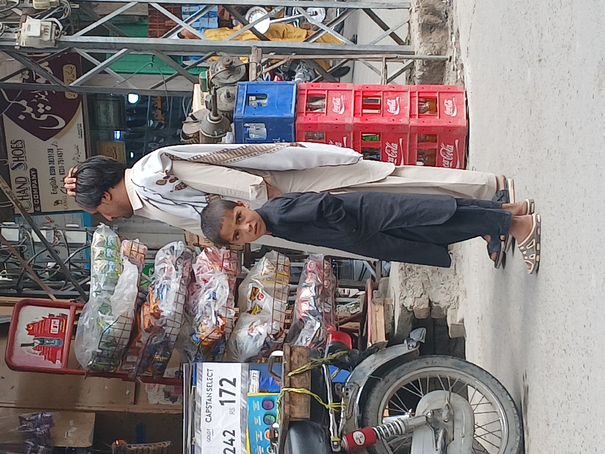 The image captures a street scene in Quetta City Tehsil, Pakistan. A man and a boy stand adjacent to a motorcycle parked in front of a shop. The man is wearing a light-colored, traditional shalwar kameez with a patterned shawl over his shoulders. The boy is dressed in a dark-colored outfit. Behind them, the shop displays snacks in clear plastic packaging. A metal rack and a sign reading "CAPSTAN SELECT" with price markings (242 and 172) are visible. The shop's signage indicates it sells "CHAND SHOES & COMPANY." Stacks of Coca-Cola crates and a blue crate are in the background. The street surface is grey and appears to be concrete or asphalt.