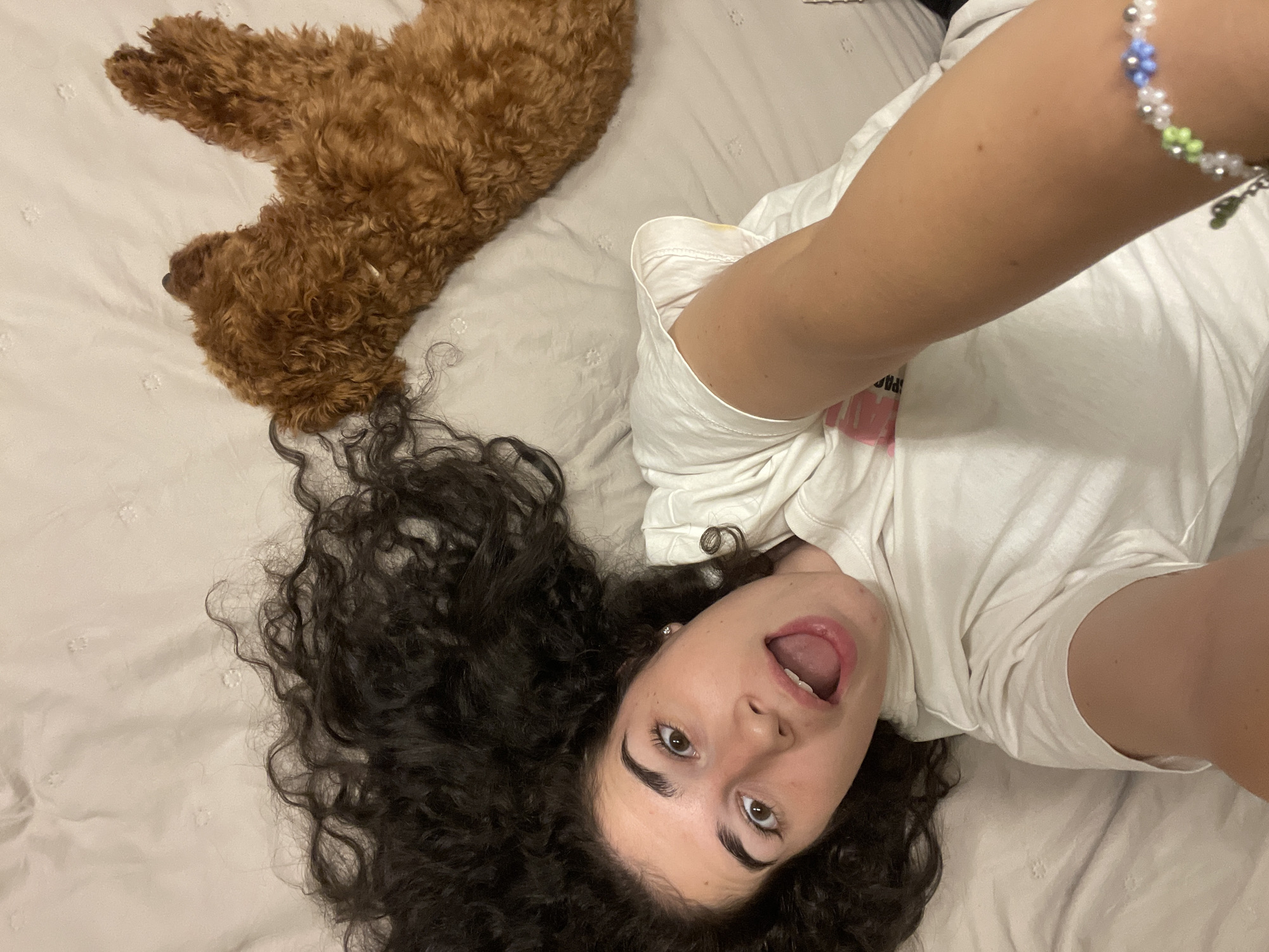 An overhead shot captures a young woman and a reddish-brown, curly-haired dog resting on a light-colored, subtly textured bed. The woman, with dark, voluminous curly hair fanned out around her head, lies on her back, looking directly up at the camera. Her expression is animated, with wide-open brown eyes and an open mouth, suggesting surprise or playfulness. She is wearing a white short-sleeved t-shirt, on which a faint, indistinct pink logo or design is partially visible near her right shoulder. A small, faint yellow stain is also visible on the shoulder area of her t-shirt. Her right arm, extended towards the camera as if taking a selfie, features a beaded bracelet composed of white, light blue, and light green flower-shaped beads, interspersed with silver beads.
Beside her, the dog, likely a poodle or doodle breed, is curled up on its side, appearing to be asleep or deeply relaxed. Its dense, reddish-brown fur is clearly visible. The setting is indoors, presumably a bedroom, illuminated by soft artificial lighting, indicating either daytime indoors or nighttime. The overall scene conveys a relaxed and intimate atmosphere, capturing a candid moment of leisure with a pet. While not discernible from the image itself, the location context for this media file is Sofia, Bulgaria.
