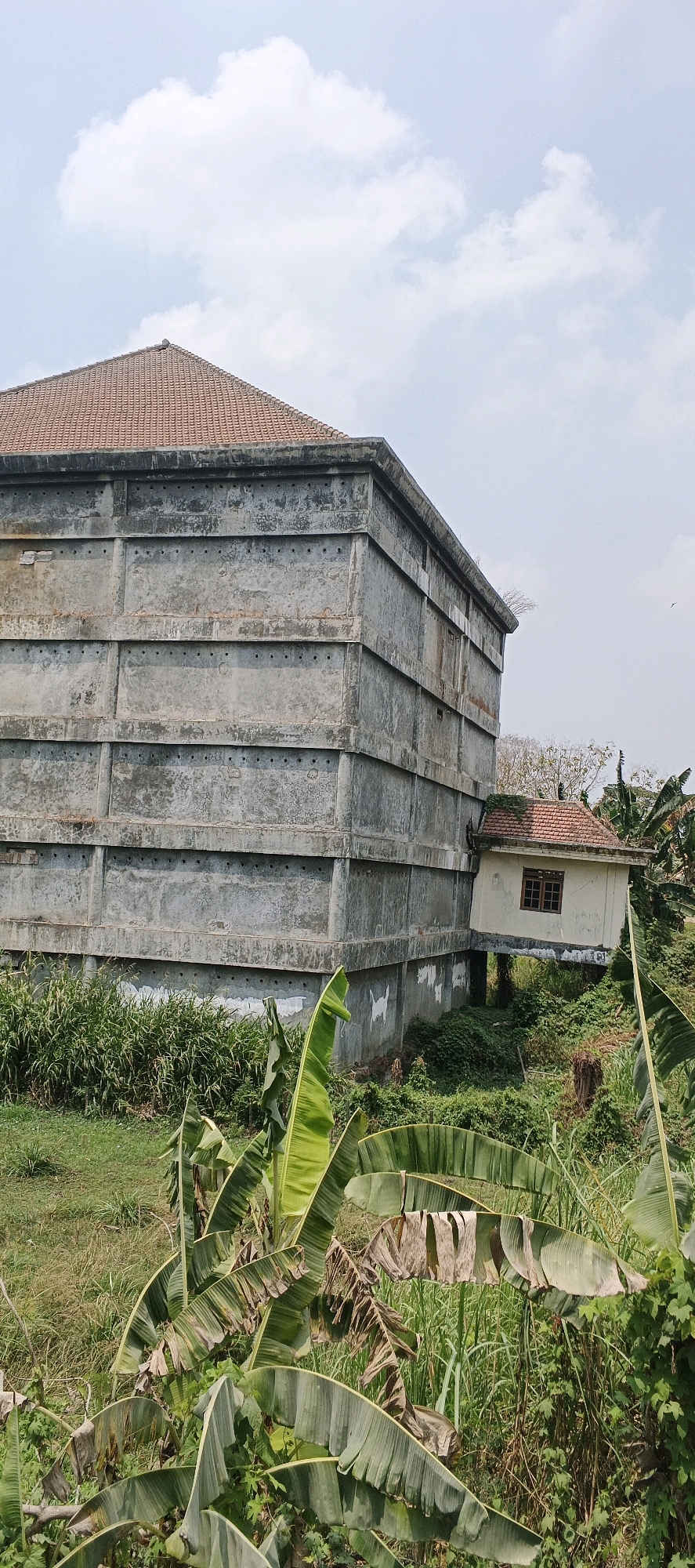 The image depicts a large, rectangular, multi-story concrete building with a red tiled roof in Laren, Indonesia. The concrete walls are rough, textured, and marked with horizontal lines and numerous small circular openings. Attached to the side of the larger building is a small, lighter-colored structure with a similar red tiled roof and a single window. Both structures are surrounded by dense green vegetation, including large-leafed plants that appear to be banana plants in the foreground. The sky is partly cloudy with a pale blue hue. The overall scene suggests an outdoor environment, possibly rural or semi-rural, with a focus on these two distinct buildings and the surrounding natural landscape.