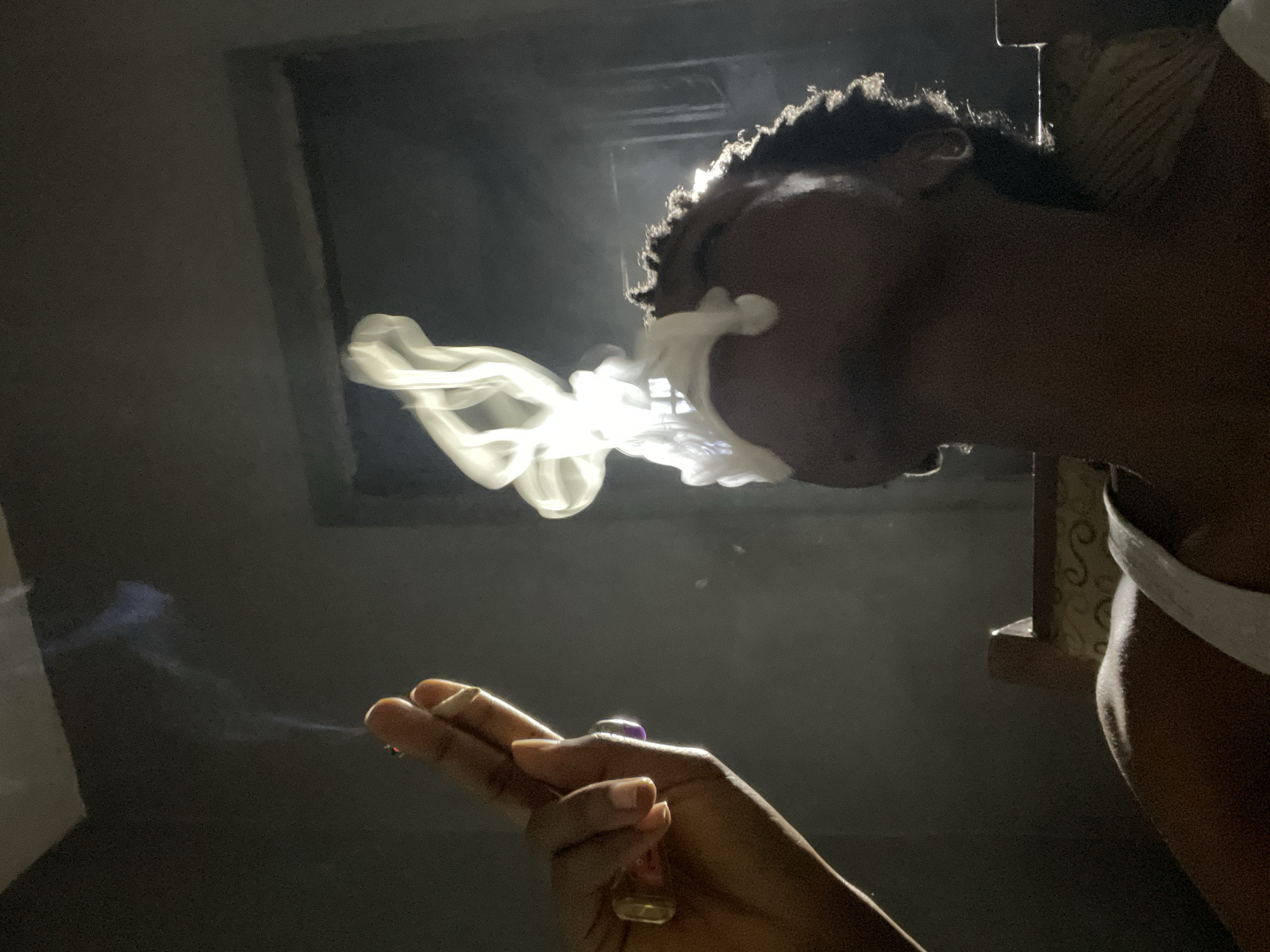 The image depicts a close-up of a person exhaling smoke. A hand holding a cigarette is visible in the left foreground, with two fingers and a thumb forming a gesture of holding the cigarette. The person exhaling the smoke is in the right foreground, with their head tilted upwards and smoke billowing out of their mouth, forming a cloud-like shape. The background is dark, with a dimly lit doorway or window visible. The overall lighting is dramatic, with strong contrasts between light and shadow. The scene appears to be indoors, likely in a dimly lit room. No specific details suggesting the location in Nkpor, Nigeria, are discernible from the image itself.