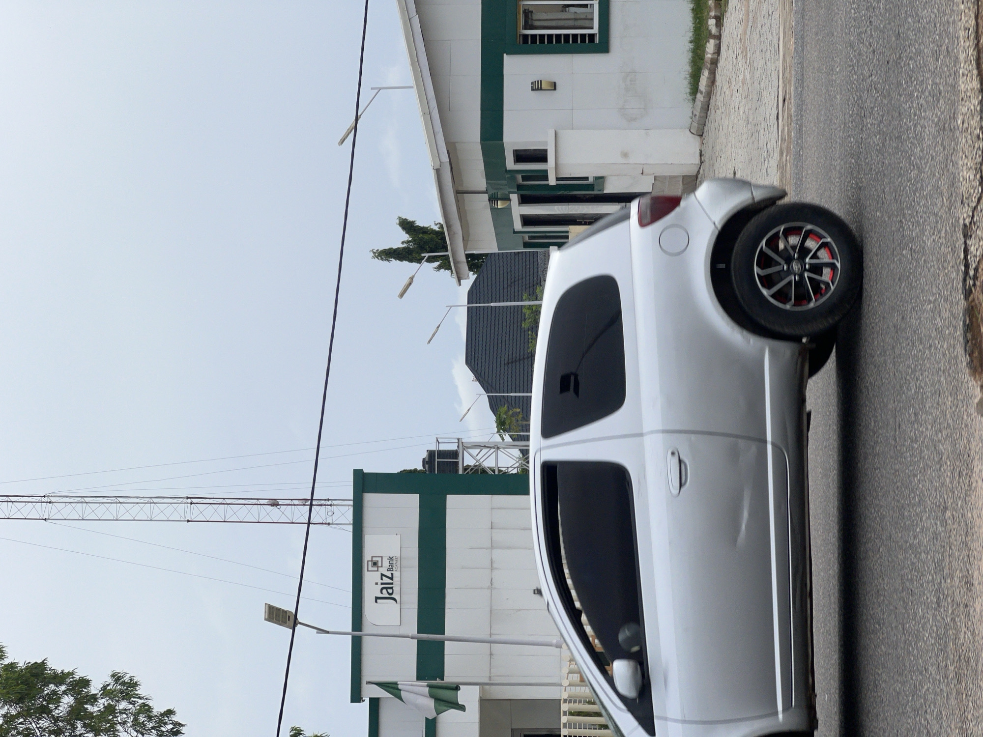 The image depicts the exterior of a Jaiz Bank branch in Jalingo, Nigeria. A silver hatchback car is driving past the bank, its side visible in the foreground. The bank building is white with green accents and features a prominent Jaiz Bank logo. A Nigerian flag is also visible on a flagpole in front of the building. In the background, a tall communication tower and several streetlights are present, set against a cloudy sky. The scene is captured during daylight hours.