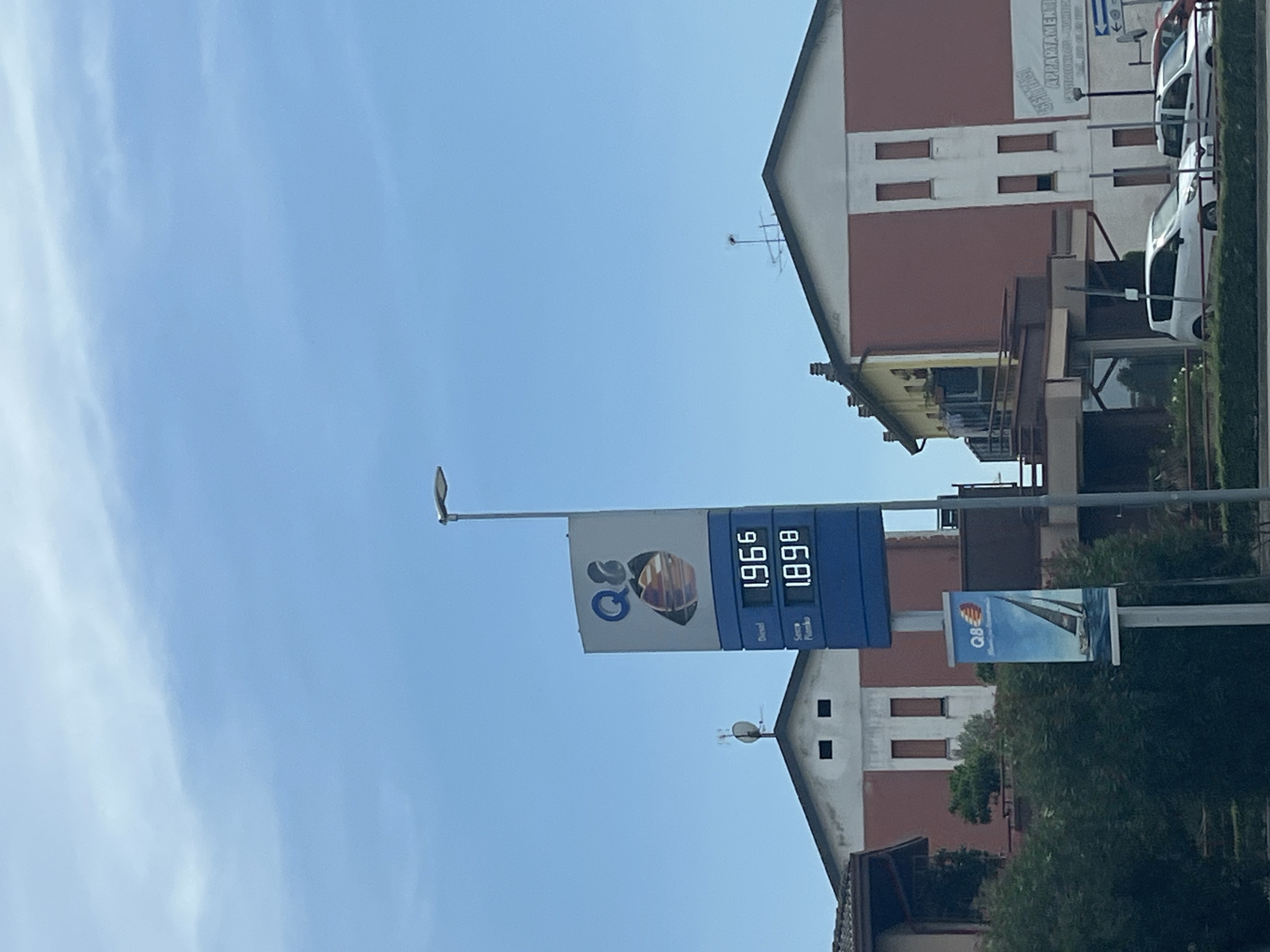 The image shows a gas station sign with prices for different types of fuel. The sign displays "Diesel" at 1.966 and "Senza Piombo" at 1.898, indicating the prices in euros per liter. Above these prices, the "Q8" logo is visible. To the left of the gas price sign, a smaller banner also features the "Q8" logo along with a sailboat. The gas station is situated in front of buildings that appear to be commercial or residential structures. The sky is clear and blue with some wispy clouds. Cars are visible parked in front of one of the buildings. The scene is captured on a sunny day. The location is identified as Castiglione delle Stiviere, Italy.
