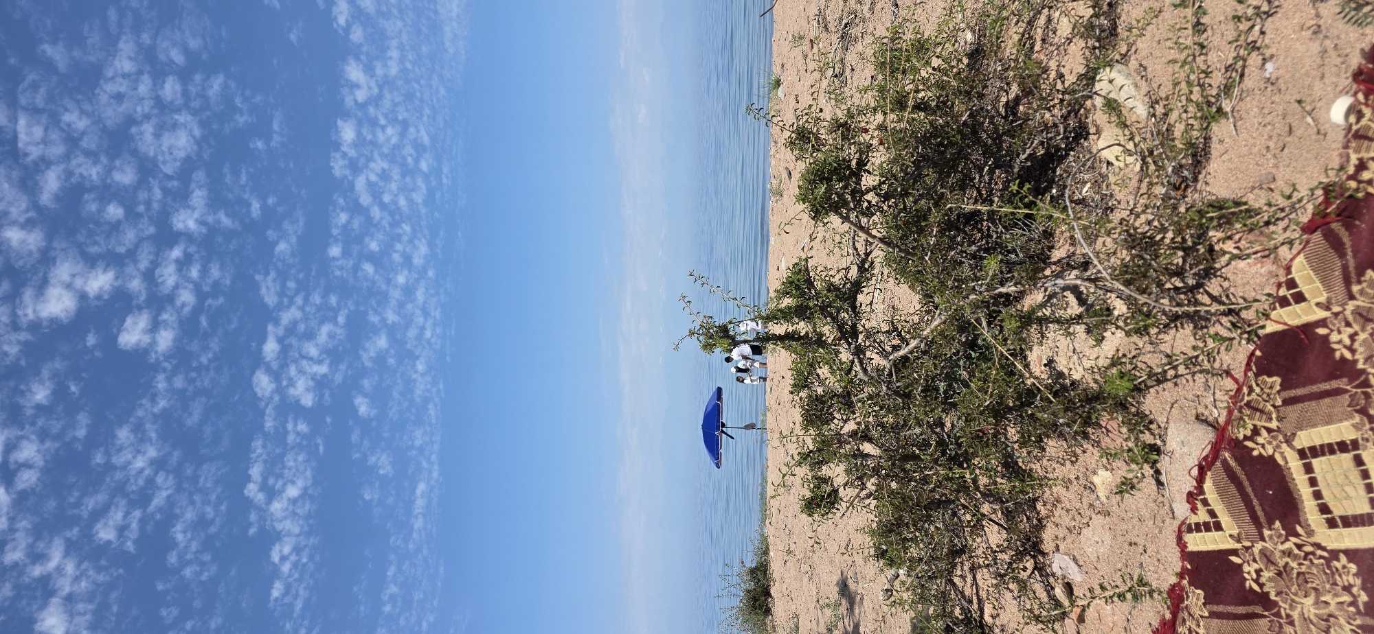 This is a vertical, medium-shot, eye-level photograph taken on a sunny day at a beach in Tosor, Kyrgyzstan, likely during daytime. The foreground is dominated by a sandy beach with sparse, dry vegetation and a portion of a patterned maroon and tan blanket. In the midground, a calm, expansive body of water stretches to the horizon, meeting a hazy blue sky filled with scattered white, cirrus clouds. A bright blue umbrella stands on the sand near the water's edge, providing shade for a small group of people. The group appears to consist of three adults and two children, all wearing white clothing. They are standing near the water, with some possibly wading or standing at the edge of the lake. The scene evokes a sense of leisure and relaxation by the water.