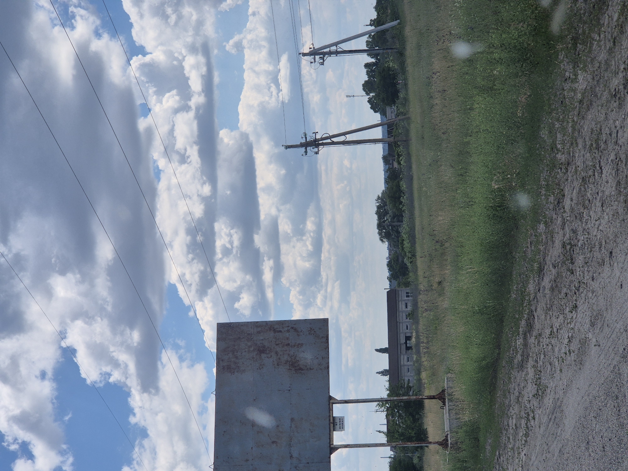The image depicts an outdoor scene on a cloudy day. In the foreground, a dirt path runs horizontally. Behind the path, a grassy field extends into the middle ground. A large, weathered metal billboard stands on the left side of the frame, supported by metal posts anchored in a concrete base. A small sign is affixed to the left support post of the billboard. In the middle ground, a long, low-slung building with a dark roof and many windows is visible, surrounded by trees. Further back, two utility poles with wires strung between them are prominent. A more distant cellular tower can be seen behind the utility poles. The sky is filled with large, white, puffy clouds against a blue sky. The overall environment suggests a rural or semi-rural setting, possibly on the outskirts of a town. The location is identified as Pidhaitsi, Ukraine.
