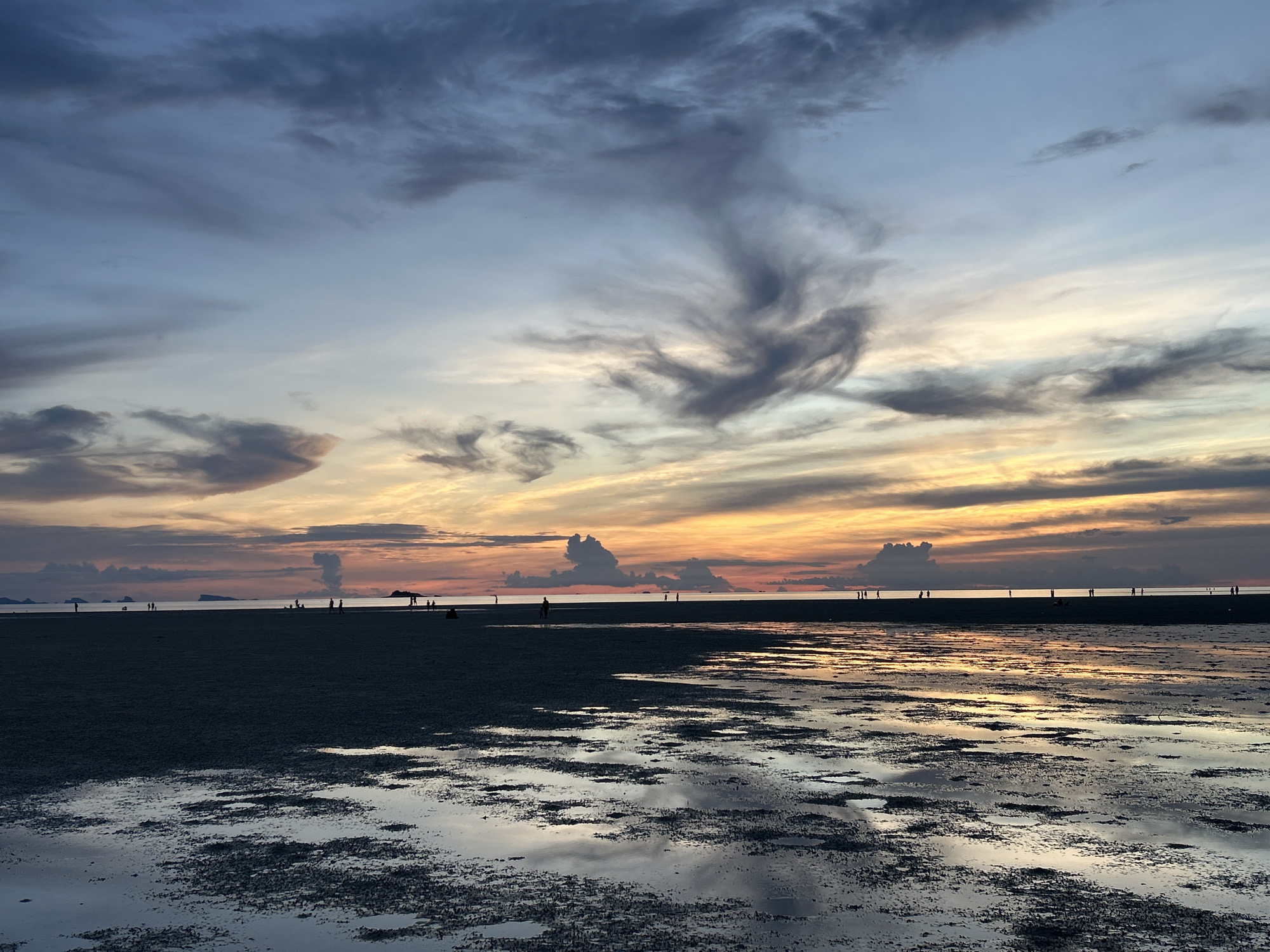 The image depicts a low tide beach scene at sunset on Koh Phangan, Thailand. The sky is a gradient of blues, grays, and oranges, with wispy clouds reflecting the sunset's hues. In the foreground, the wet sand is covered with shallow pools of water, creating a mirror-like surface that reflects the sky and clouds. A dark sandbar stretches across the middle ground, populated by several silhouetted figures of people walking and standing, some engaged in activity. In the far distance, the horizon shows a faint outline of land or islands, with more silhouetted figures on what appears to be a distant shoreline. The overall atmosphere is serene and tranquil, capturing the beauty of a tropical sunset over the ocean.