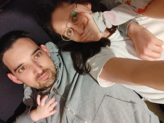 The image, taken indoors in Tel Aviv-Yafo, Israel, presents an overhead view of two adults and a young child lying closely together on a dark, soft surface, likely a bed or couch. An adult male is positioned on the lower left, featuring short dark hair, a light beard, and light-colored eyes, looking directly at the camera. He wears a grey zippered hooded jacket. To his right, an adult female with long dark hair and round gold-rimmed glasses looks towards the camera, her left hand partially covering her lower face. She wears a light short-sleeved top layered over a multi-colored pastel striped long-sleeved garment. A small hand, consistent with that of an infant or young child, rests on the male's chest, over his jacket. The specific indoor location within Tel Aviv-Yafo is not discernible from visual cues.