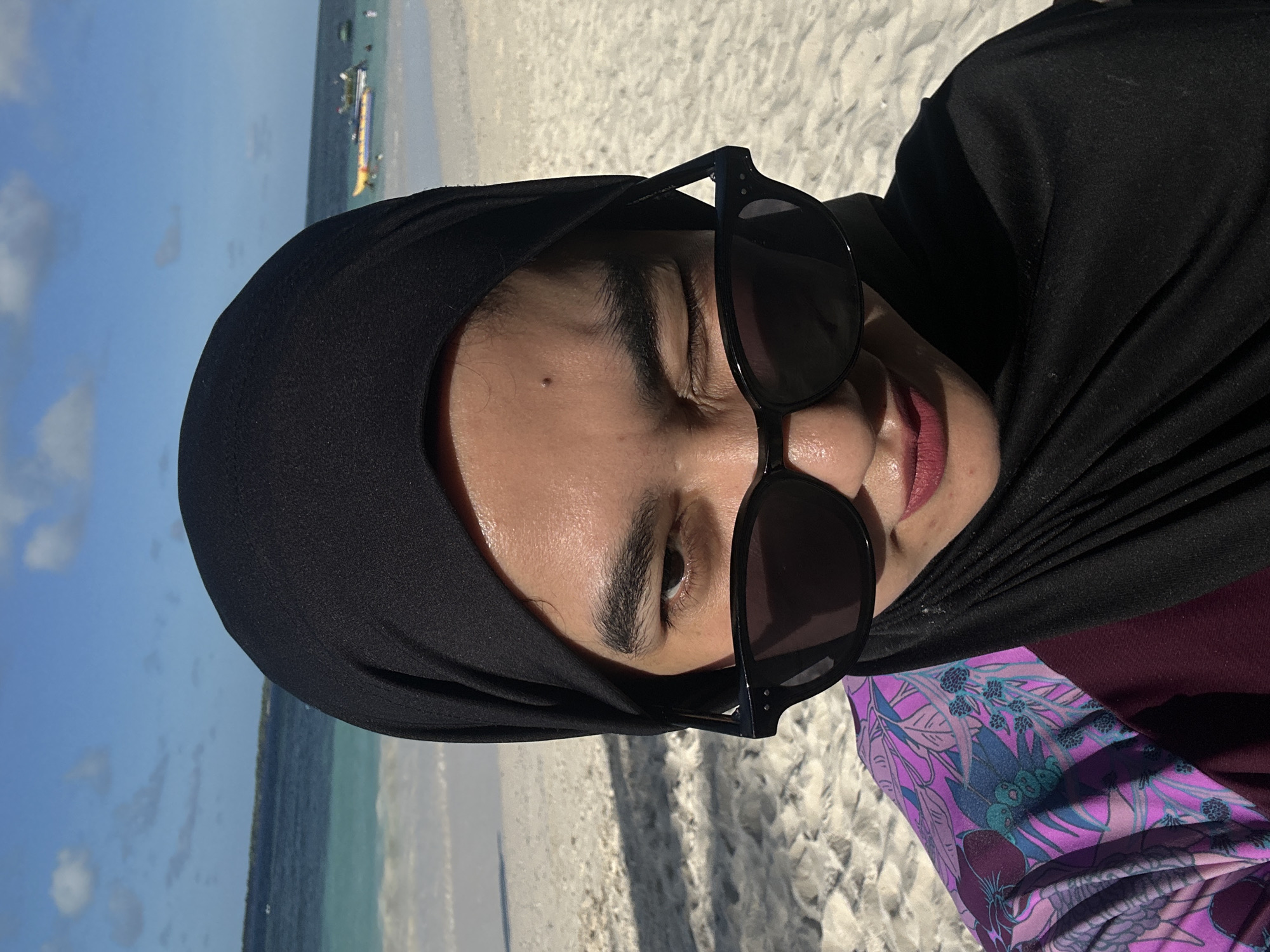A woman wearing a black hijab and sunglasses winks at the camera while on a sandy beach. She is wearing a floral-patterned top. The ocean and sky are visible in the background, with a few boats in the distance. The location is identified as the city of A R A, Indonesia.