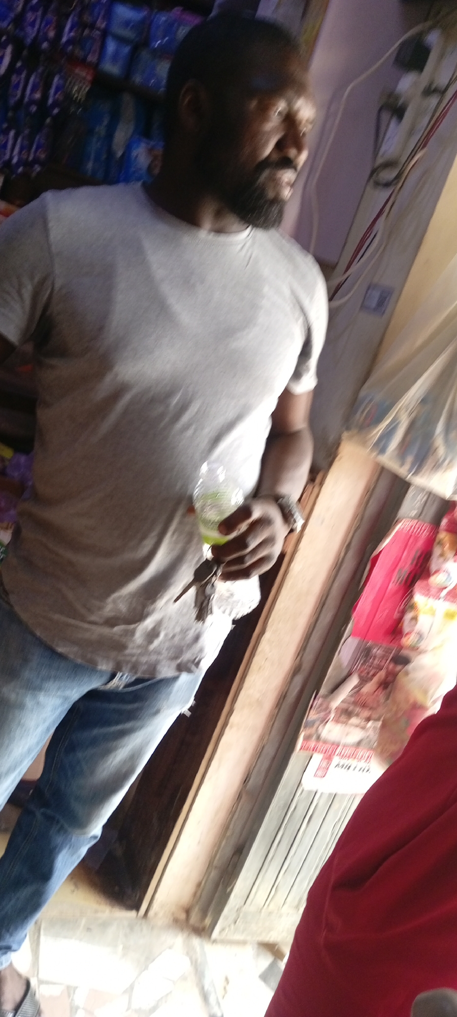 An indoor, slightly tilted medium shot captures a man standing in what appears to be a small shop or store in Gayawa, Nigeria. The man has a dark complexion, a beard, and short dark hair. He is wearing a light grey short-sleeved t-shirt, blue jeans, and a watch on his left wrist.
In his left hand, he holds a clear plastic bottle containing a bright green liquid, likely a beverage, along with a small bunch of keys. He is looking towards his right, out of the frame. His expression is calm.
The background is somewhat blurred but shows shelves stacked with various items, suggesting merchandise in a retail setting. On the far right side of the image, there is a wooden structure, possibly a doorframe or counter. Beyond this, a person in a red garment is partially visible. Further back, a red and white poster or packaging can be seen on a wall or surface, with the word "MILO" faintly visible in white text on a red background. The floor beneath him appears to be tiled. The lighting is bright, indicating it is likely daytime.