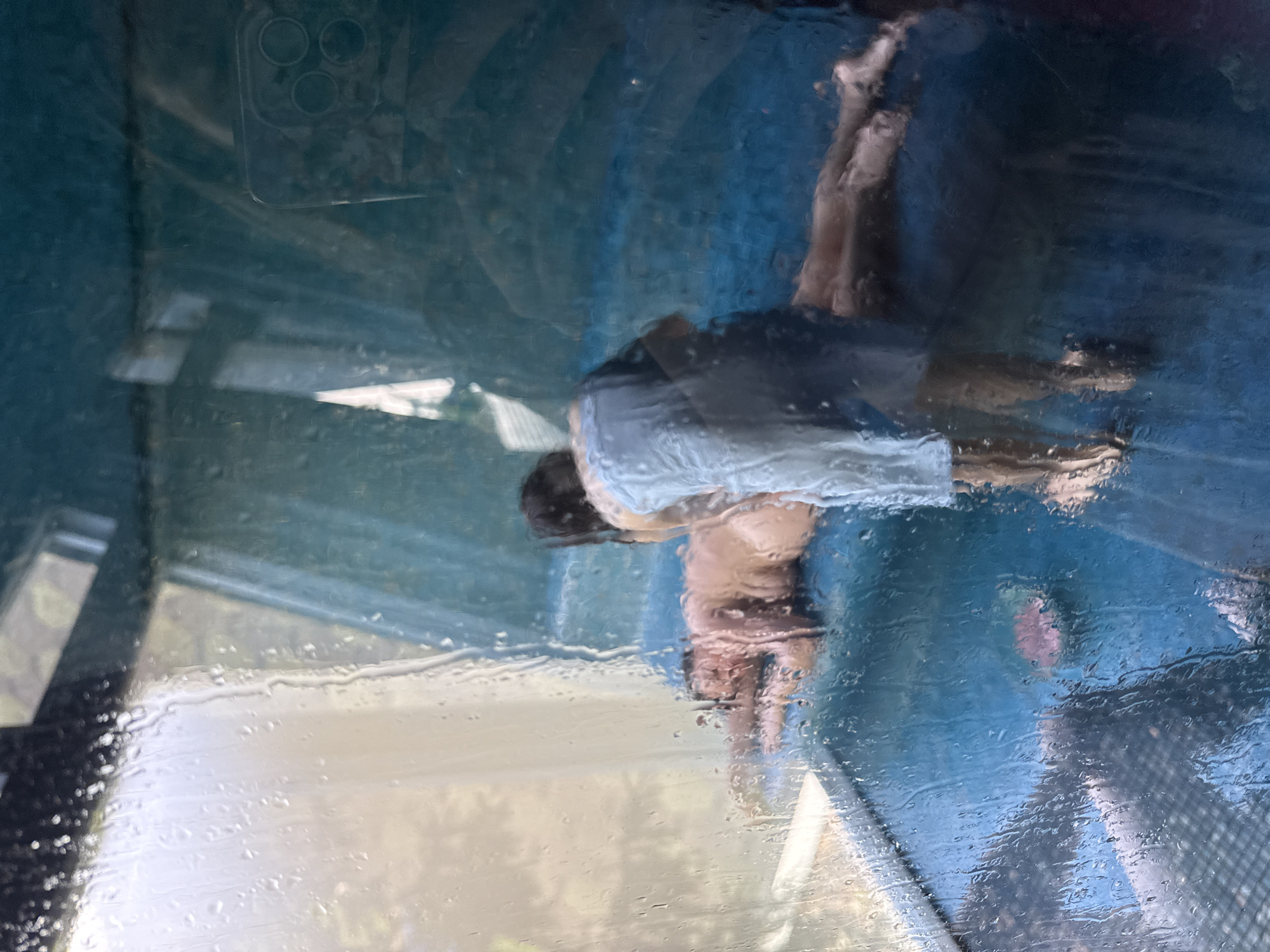 The image, shot through a rain-streaked window, shows a partial reflection of a person. The individual, wearing what appears to be a light-colored shirt and dark shorts, is partially obscured by the distorted reflection. The camera of the device taking the picture is visible in the upper left corner of the frame. The background shows some indistinct shapes and colors, suggesting an outdoor setting. There is no evidence of the city of Chornoye, Russia, in the image.