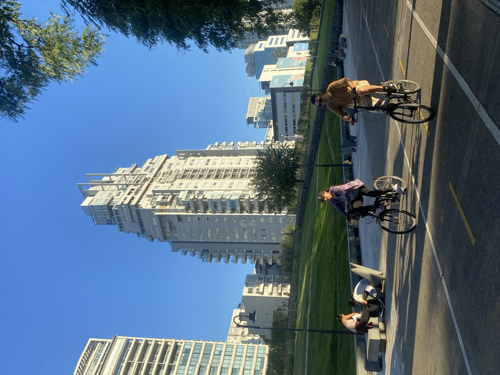The image depicts a sunny outdoor scene in Buenos Aires, Argentina, featuring cyclists and pedestrians against a backdrop of modern architecture.
Two individuals are riding bicycles on a paved path marked with yellow lines. One cyclist, a man, wears a brown shirt and light shorts. The other, a woman, wears a purple patterned jacket and black leggings.
To the side of the path, a grassy area is adjacent to a low concrete wall where three people are sitting on benches. A streetlight stands nearby.
Behind the park scene, several tall, modern buildings fill the skyline, identifiable as part of the Puerto Madero neighborhood. The clear blue sky indicates a bright, sunny day.