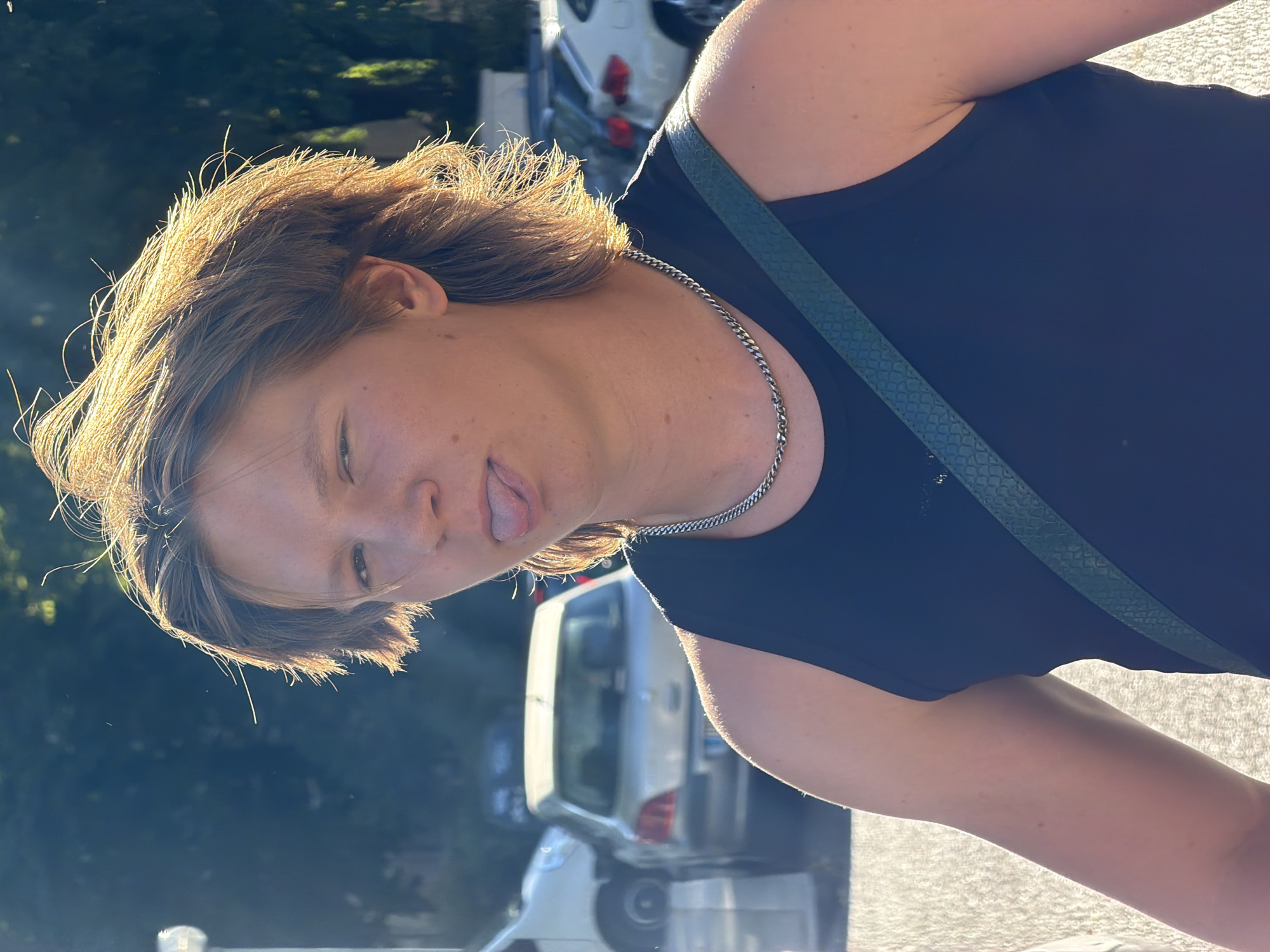 The image is an outdoor, eye-level, medium shot of a person with light brown hair wearing a black tank top and a silver chain necklace. The person is positioned in the center of the frame, with their head tilted to the side and their tongue sticking out. The background is blurred and consists of trees, parked cars, and sunlight creating lens flares. The sunlight suggests it is daytime, likely late afternoon given the warm tones. The scene appears to be a parking lot or street area, possibly in a residential or commercial district. The overall mood of the image seems playful and candid. No visible text is present in the image. The context provided suggests the location is Lyon, France.