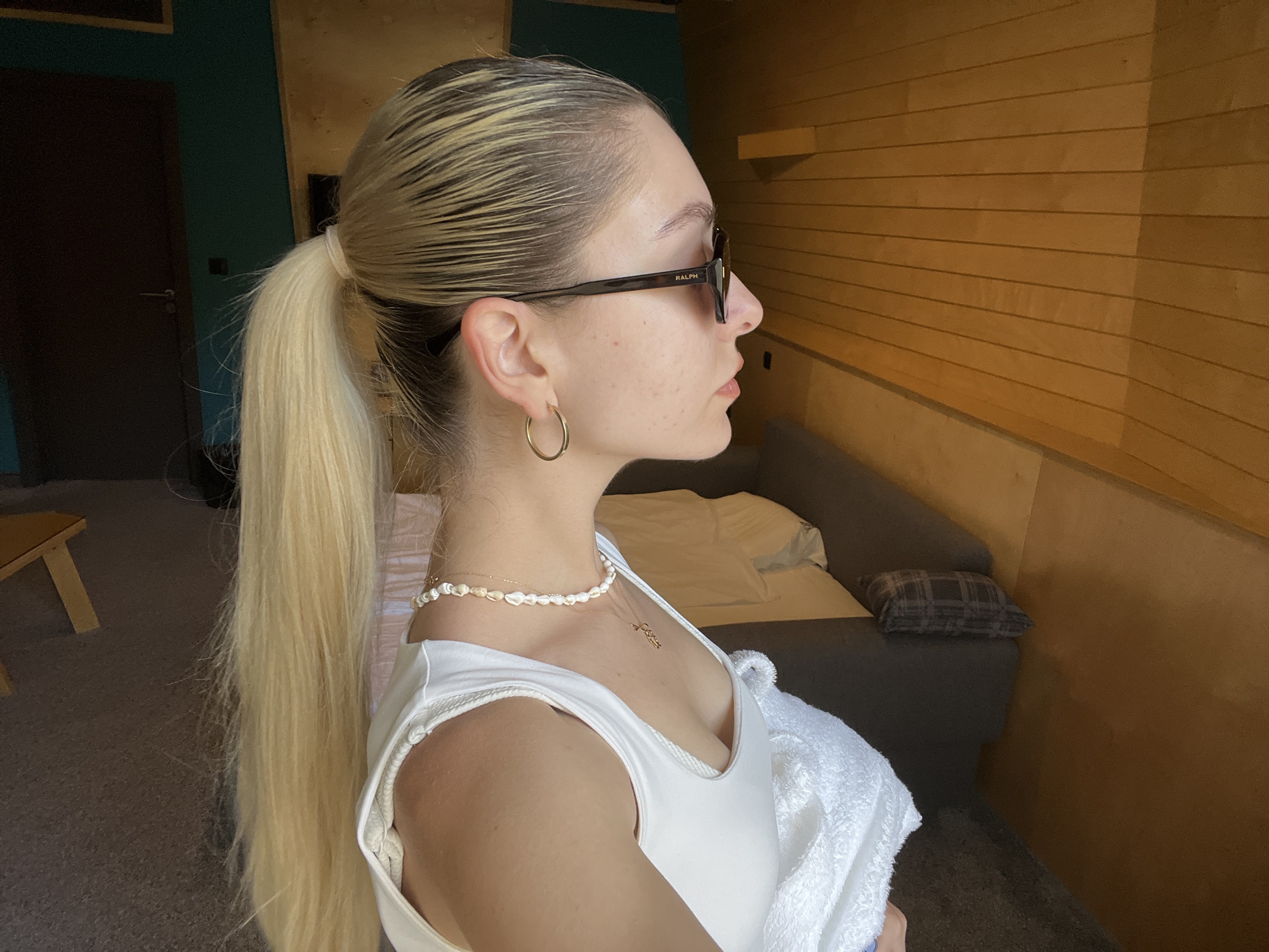 A fair-skinned person with blonde hair pulled back in a ponytail is viewed in profile. They are wearing sunglasses and a white tank top, with a pearl necklace and a gold hoop earring. Behind the person is a room with wood paneling and a gray couch. The city and exact location are unknown.