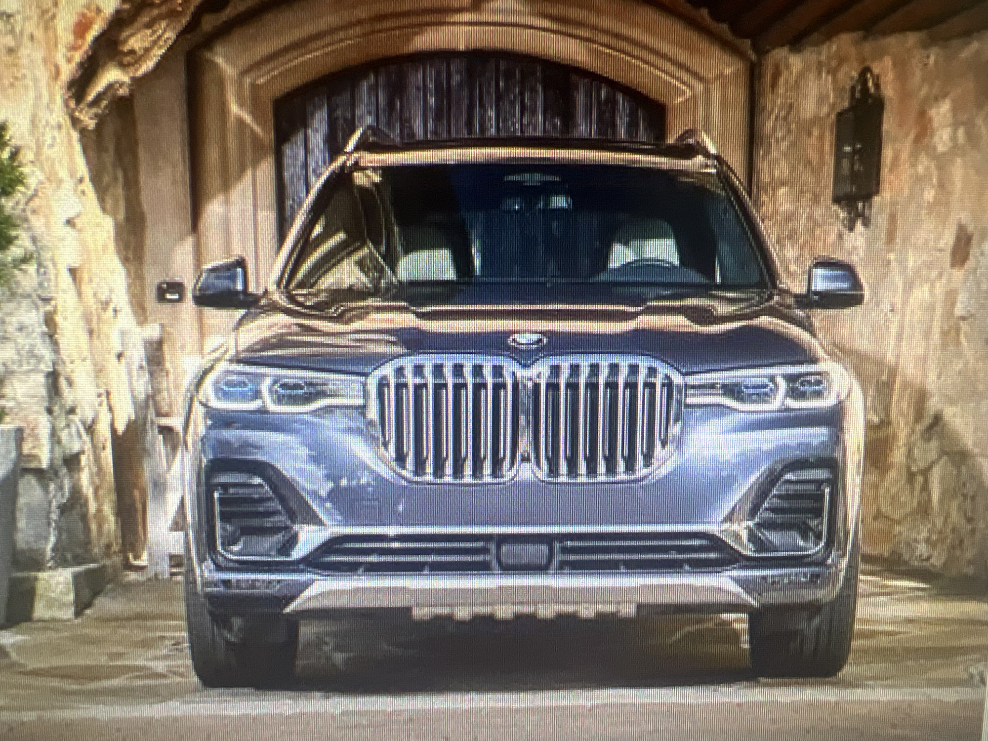 A front-facing, medium shot captures a dark blue BMW X7 SUV parked in a stone-clad entryway, possibly a garage or driveway, with a large wooden door in the background. The SUV's headlights are on, emitting a white glow. The vehicle's grille is prominent, featuring vertical chrome slats and the BMW logo. The surrounding walls are textured stone, with a dark-colored light fixture mounted on the right wall. The ground is paved with stone or concrete. The overall impression is one of a luxury vehicle in a well-maintained, possibly upscale, setting. The image does not contain any identifiable landmarks that would pinpoint its location in Cheb, Czechia.