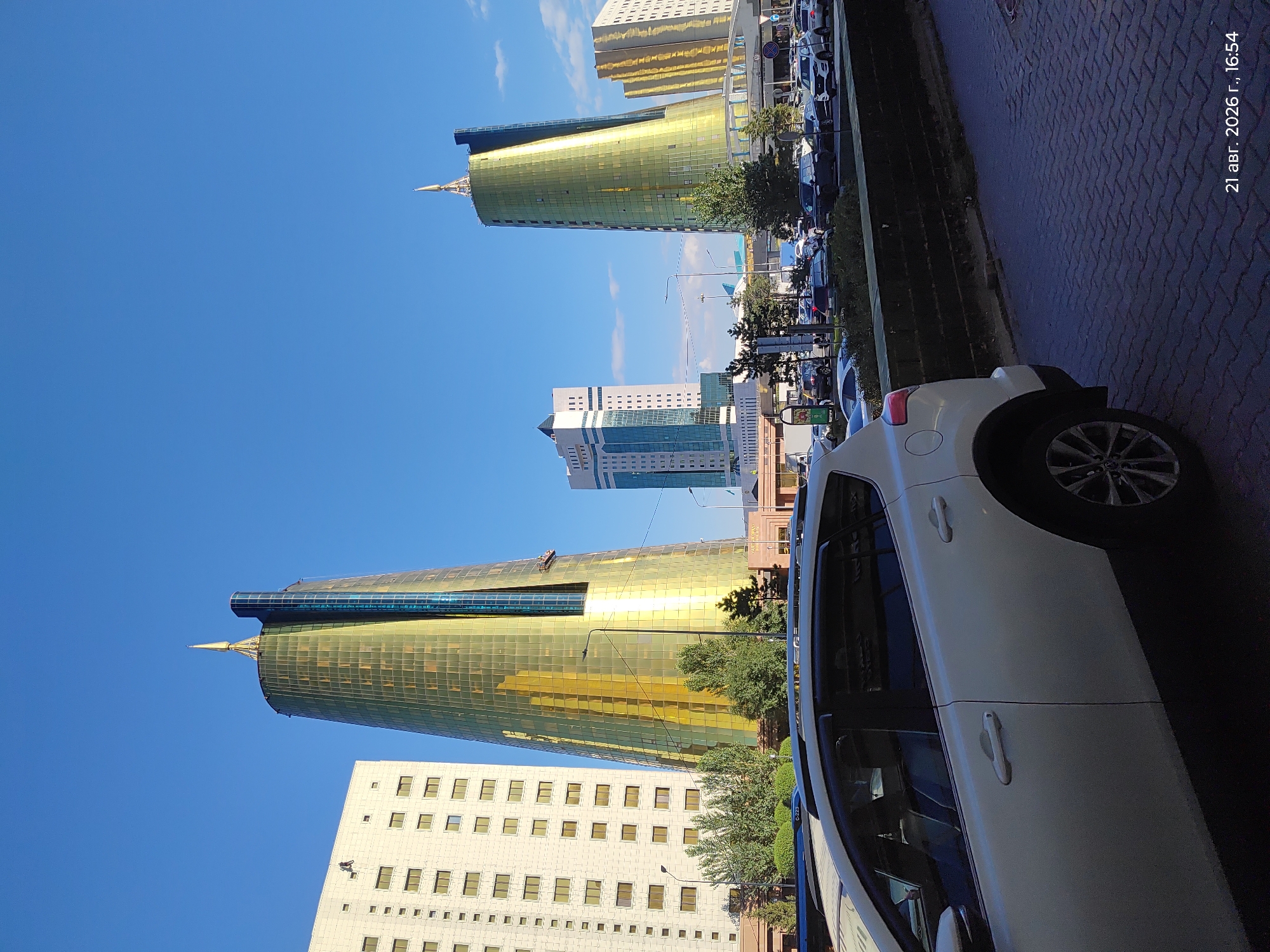 The image depicts a street scene in Astana, Kazakhstan, on August 21, 2026, at 16:54. The foreground is dominated by the rear passenger-side view of a white SUV. In the background, several prominent skyscrapers are visible under a clear blue sky. The most striking is a tall, slender, golden-colored skyscraper with a pointed spire. To its left is a rectangular, light-colored building with numerous windows. Further to the right, another golden skyscraper, similar in design but shorter than the first, stands adjacent to a white, rectangular building with a patterned facade. A row of trees and bushes separates the street from the buildings. Several other cars are parked and driving on the street. The sidewalk in the foreground is paved with dark, rectangular stones. A street lamp is visible between the white SUV and the golden skyscraper.