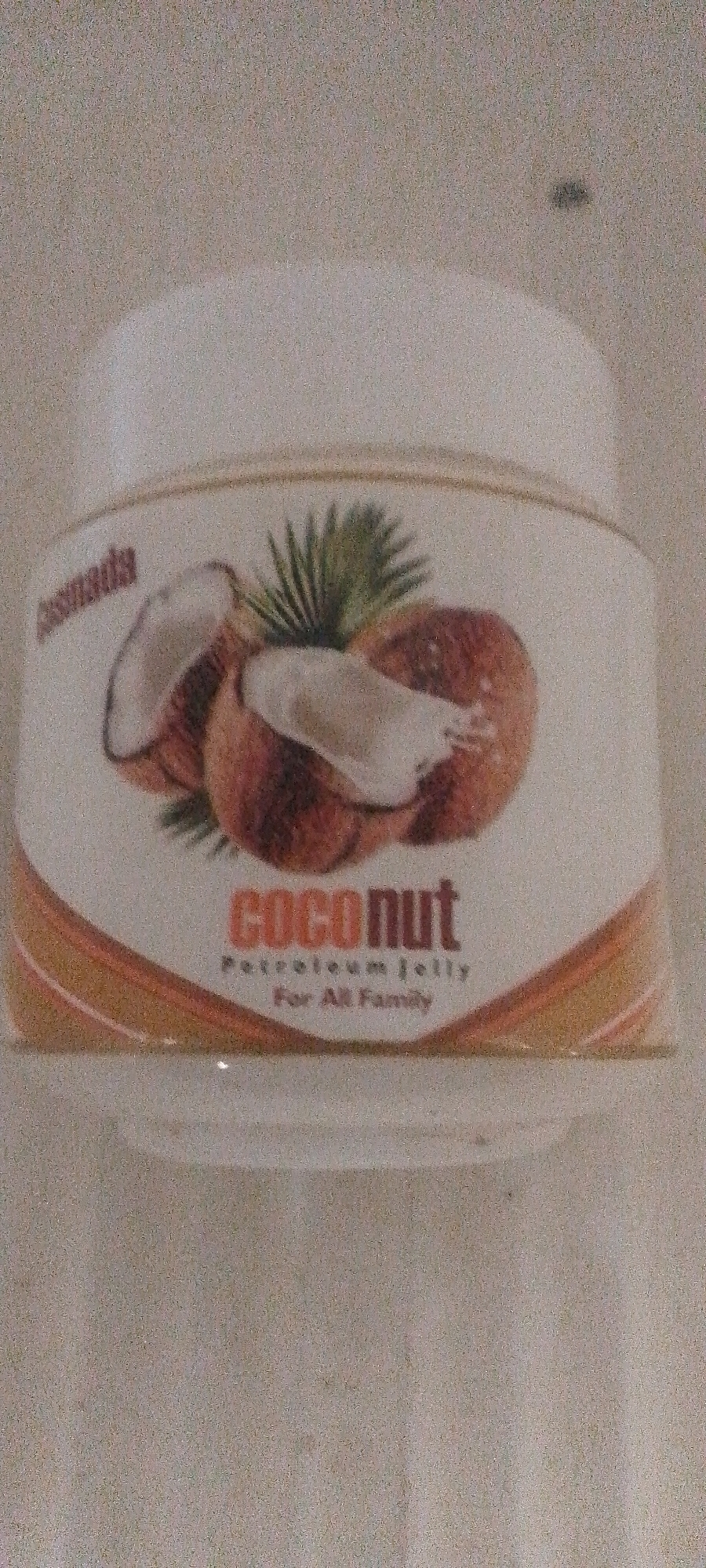 A close-up shot of a jar of petroleum jelly with a coconut theme is captured. The jar has a white lid and a label with a graphic of two halves of a coconut and a palm frond. The text on the label reads "cosmada" in a smaller font on the left, and "coconut" in larger letters in the center. Below "coconut," smaller text reads "Petroleum Jelly" and "For All Family." The background is a muted, textured surface, possibly a wall or countertop, and there are some digital artifacts or graininess throughout the image. The lighting is diffused. The image appears to have been taken indoors. The image does not contain any information about Jalingo, Nigeria.