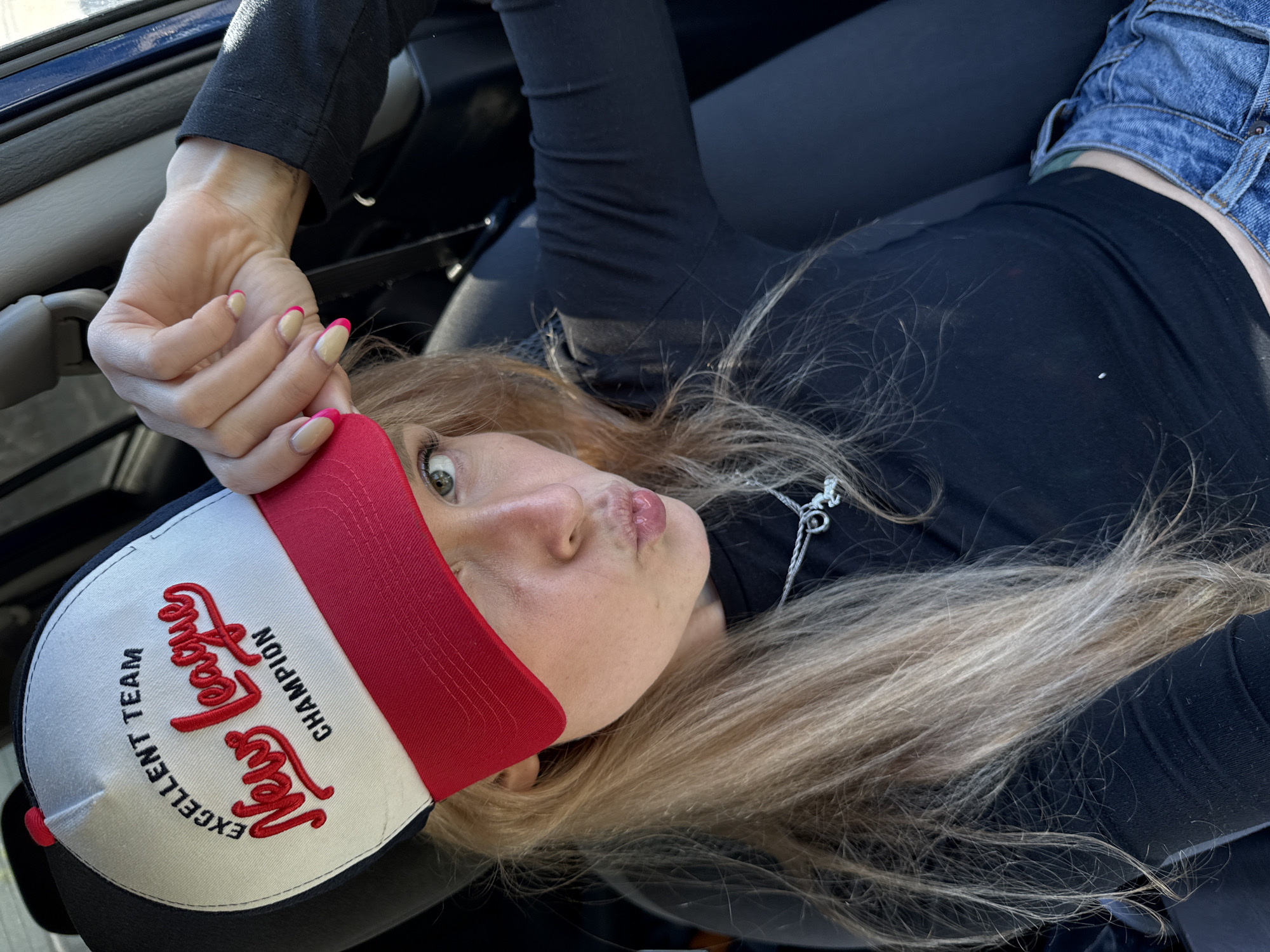 A fair-skinned, adult female with long, blonde hair is lying supine in a vehicle. She is wearing a black, long-sleeved shirt and denim shorts. A baseball cap with "Excellent Team New League Champion" embroidered on it obscures part of her forehead. Her nails are manicured with pink tips. The interior of the vehicle is visible, with a blue dashboard and dark seating. The location is unknown; there is no identifiable information to suggest it is Chaikovsky, Russia.