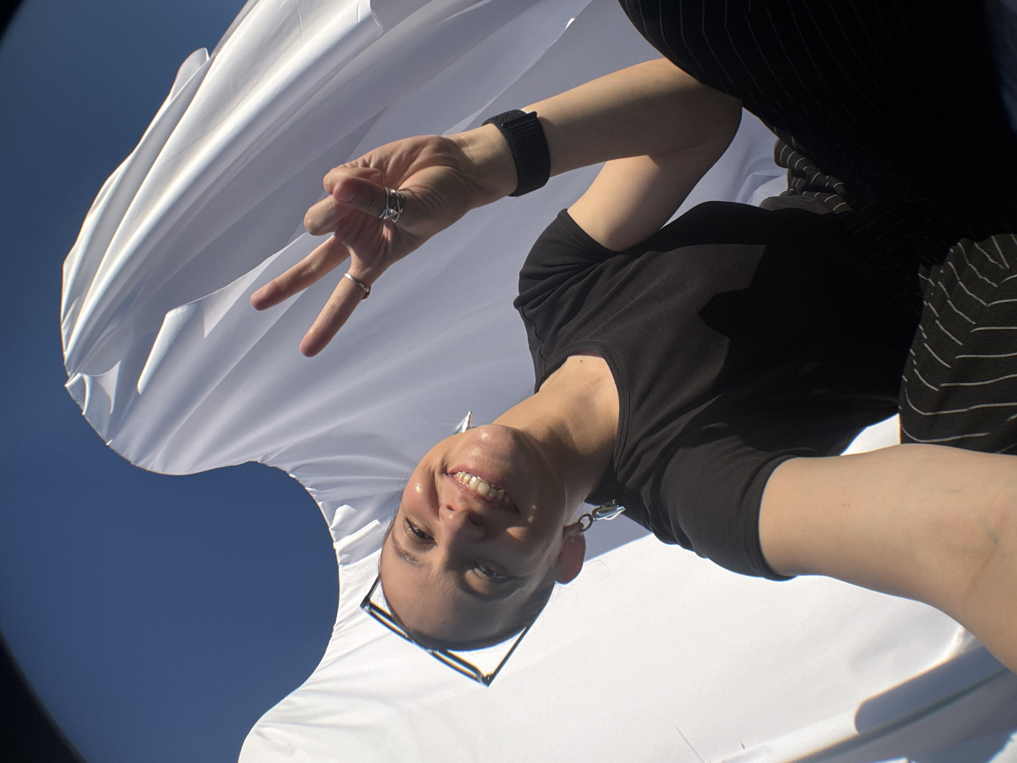 In a low-angle, wide-lens photograph taken outdoors, a person with short dark hair, wearing glasses perched on their head, smiles broadly at the camera. They are wearing a black t-shirt and striped pants. Their left hand is raised, fingers forming a peace sign, adorned with two silver rings. A black wristband is visible on their left wrist. The subject is positioned against a backdrop of flowing white fabric, possibly a tent or draped material, with a clear blue sky visible above and to the left. The lighting suggests a sunny day. The location is identified as Ольховка, Russia.