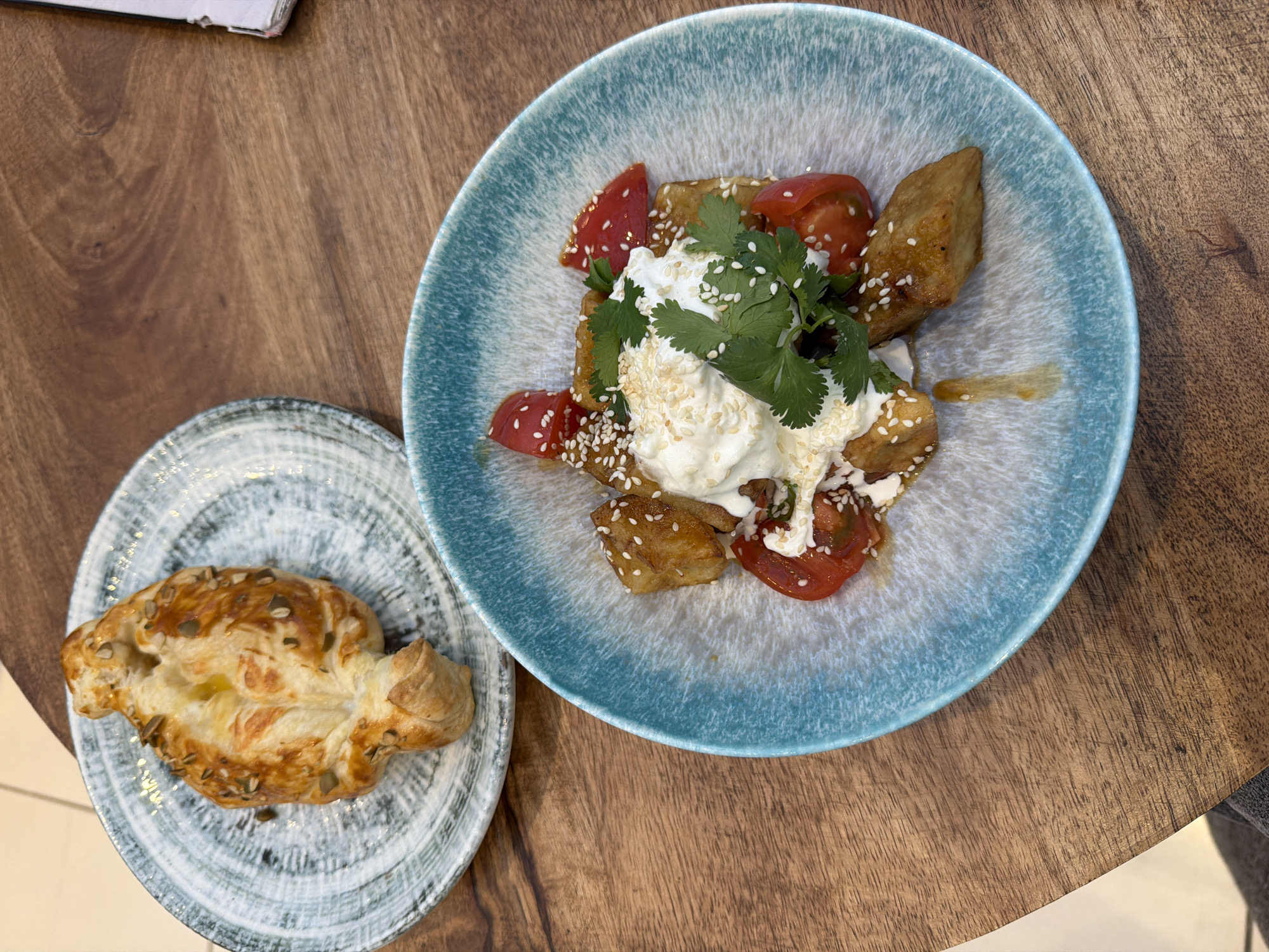 An overhead shot captures two distinct dishes presented on a textured wooden table, suggesting a meal or snack in a casual dining setting in Khimki, Russia.
The focal point is a larger, round ceramic plate with a speckled blue and white glaze, holding a vibrant dish. This main dish consists of golden-brown, fried pieces, likely eggplant or similar vegetable, arranged with bright red, quartered tomatoes. A generous dollop of white, creamy sauce or yogurt sits in the center, garnished with fresh green cilantro sprigs and a liberal sprinkling of white sesame seeds. A faint amber drizzle, possibly a sauce or oil, is visible on the plate's surface.
Adjacent to the larger plate, on a smaller, similarly textured gray and white ceramic plate, rests a single, golden-brown pastry. This pastry appears to be made from puff pastry, with a flaky, airy texture and is topped with scattered seeds, likely pumpkin and/or sesame seeds, adding to its rustic appeal.
The wooden table features a rich, warm brown hue with visible grain patterns, indicating natural wood. In the upper left corner, a portion of an open book or menu is visible, hinting at a cafe or restaurant environment. No people are explicitly visible, though the presence of dishes implies a diner. The lighting suggests it is daytime. No discernible text is visible on the dishes or background elements.