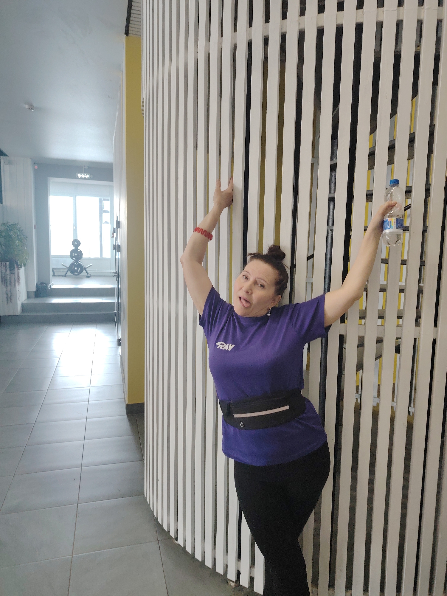 A woman is leaning against a curved white slat wall in a hallway. She is wearing a purple t-shirt, black leggings, and a black fanny pack. She holds a plastic water bottle in her right hand and her tongue is sticking out. In the background, a hallway leads to a window with exercise equipment visible. The location is Sarapul, Russia.